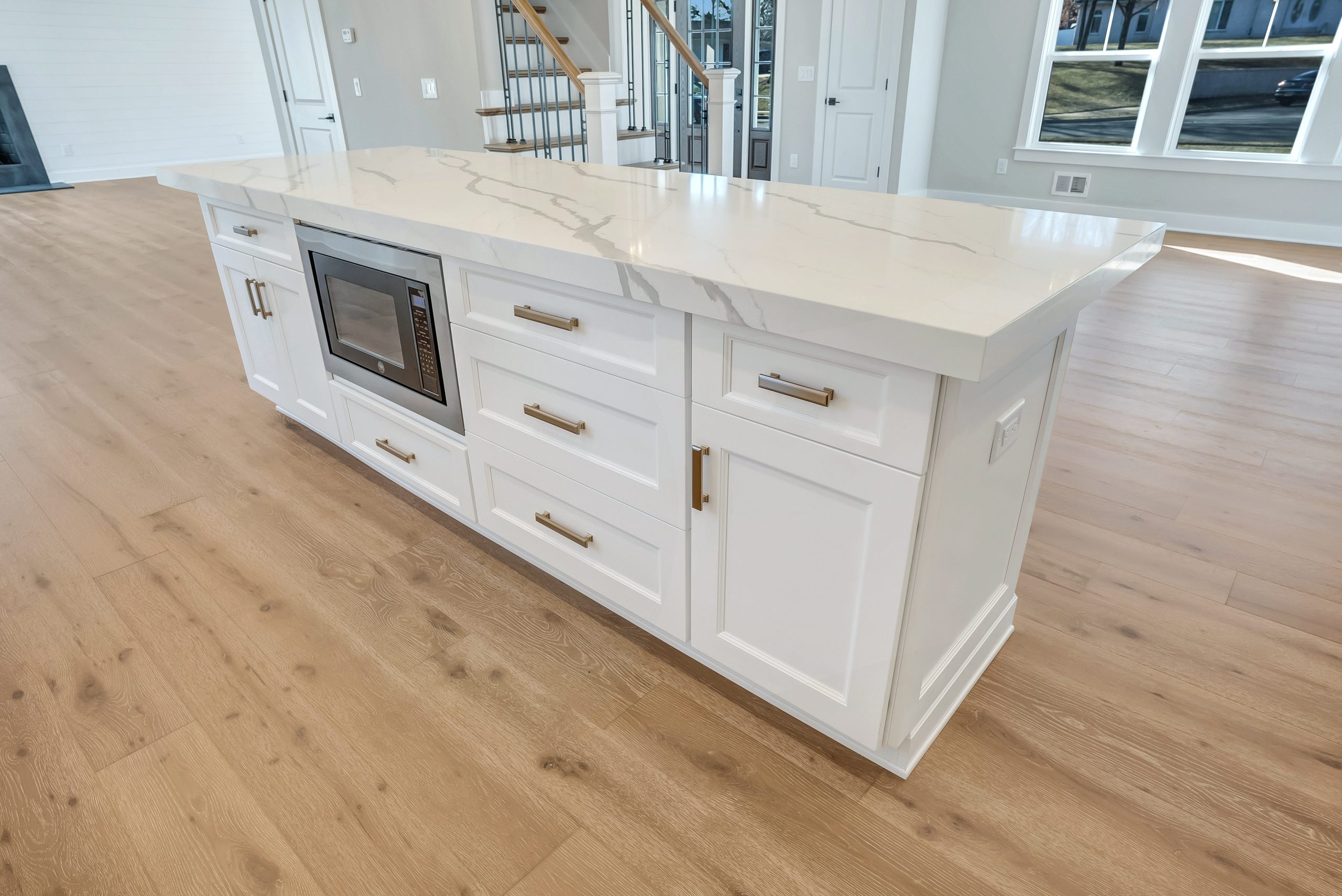 A modern kitchen island with a white marble countertop, built-in microwave, white cabinets with gold handles, and light wood flooring in a bright, spacious room.