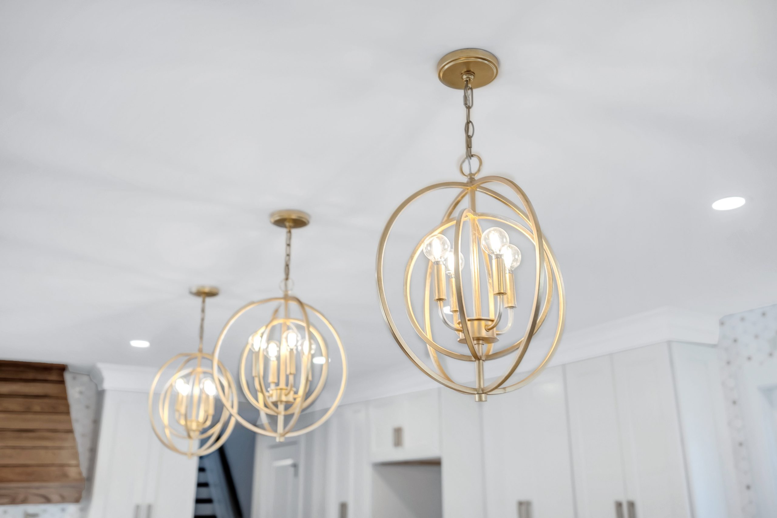 Three modern gold spherical pendant lights with exposed bulbs hang from a white ceiling in a bright, contemporary room with white cabinets and recessed lighting.