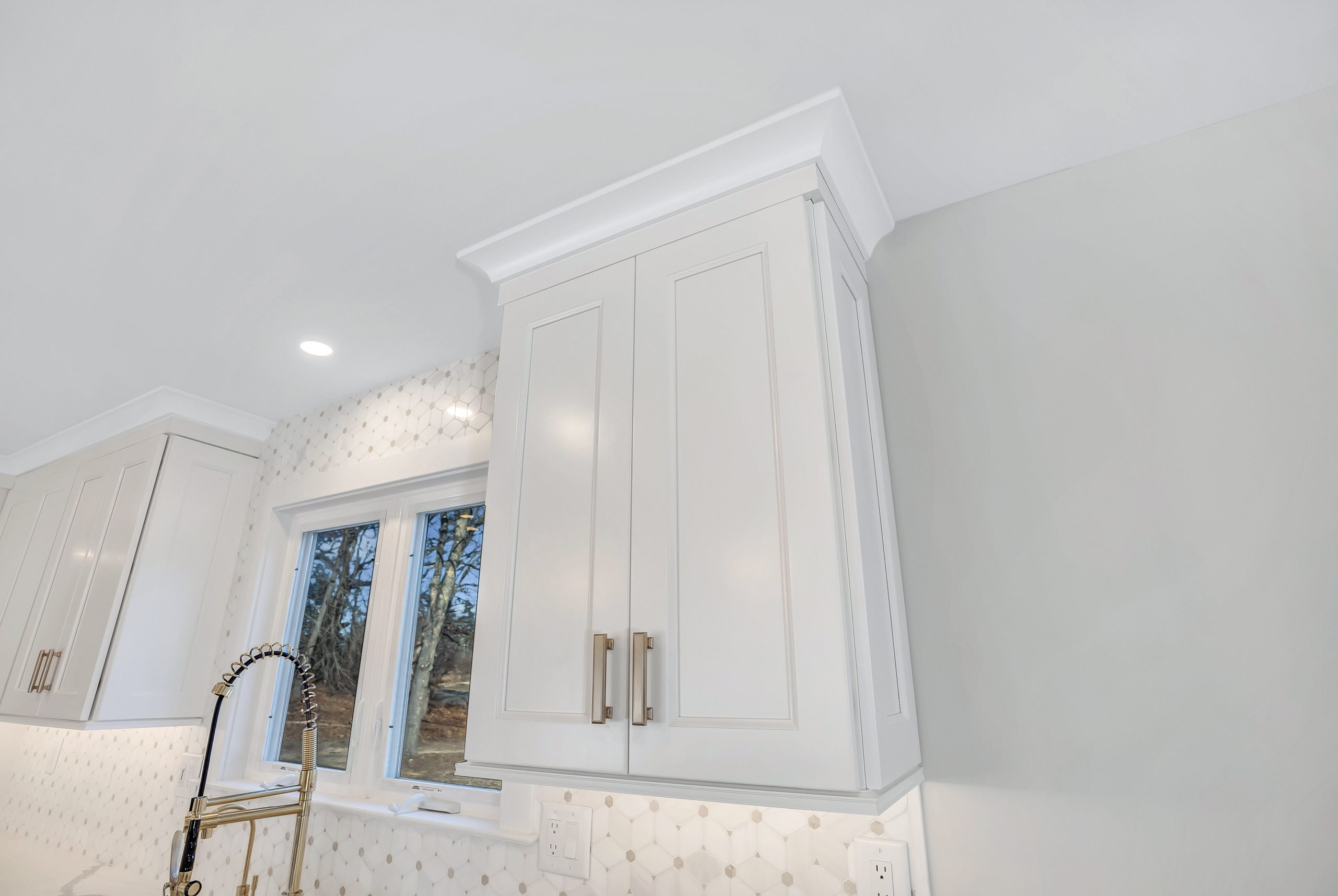 White kitchen cabinets with silver handles, a window with a view of trees, a modern faucet, and a white geometric tile backsplash under bright overhead lighting.