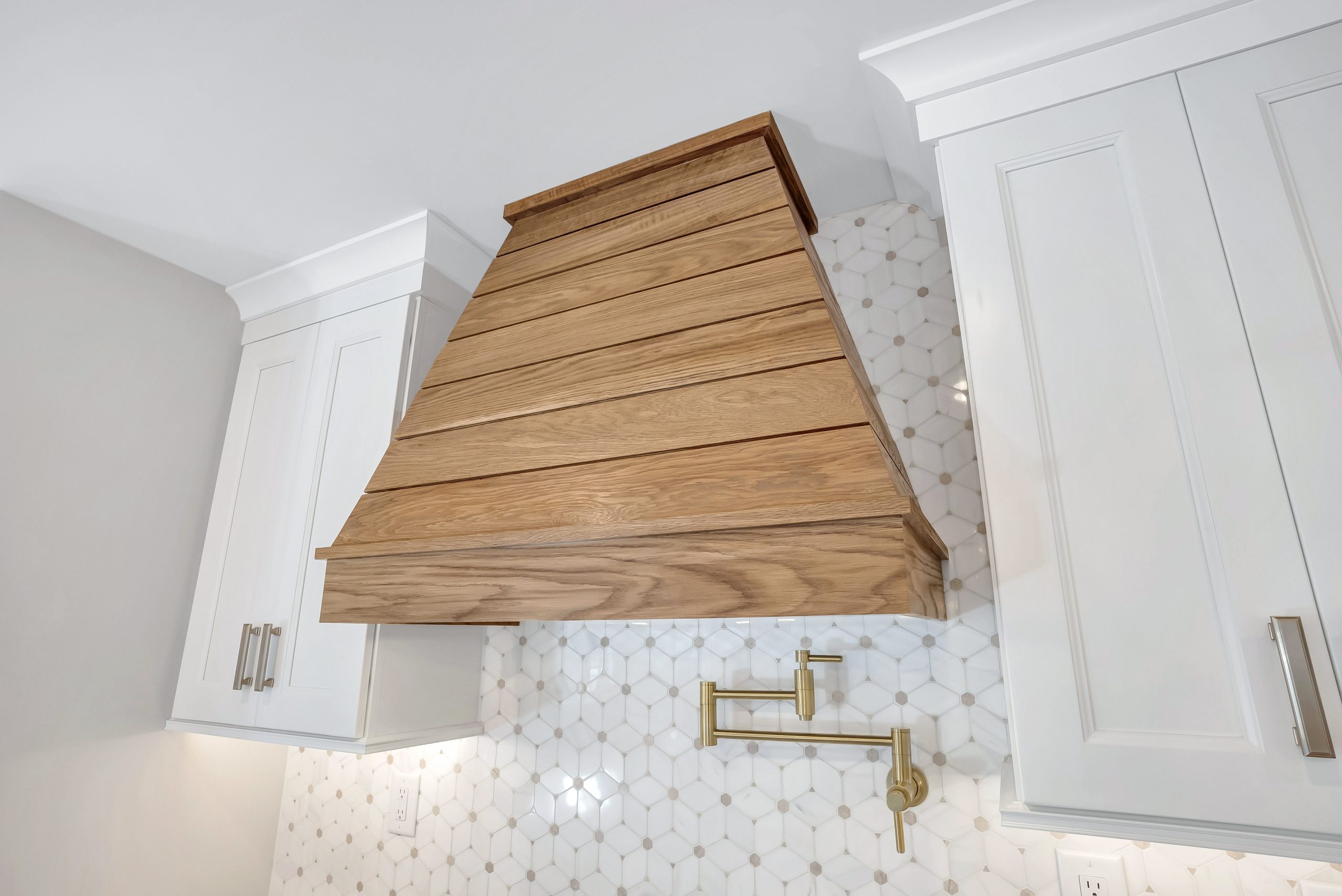 Wooden kitchen range hood with a slatted design, flanked by white cabinets. The backsplash features white hexagonal tiles, and there is a gold pot filler faucet mounted on the wall.