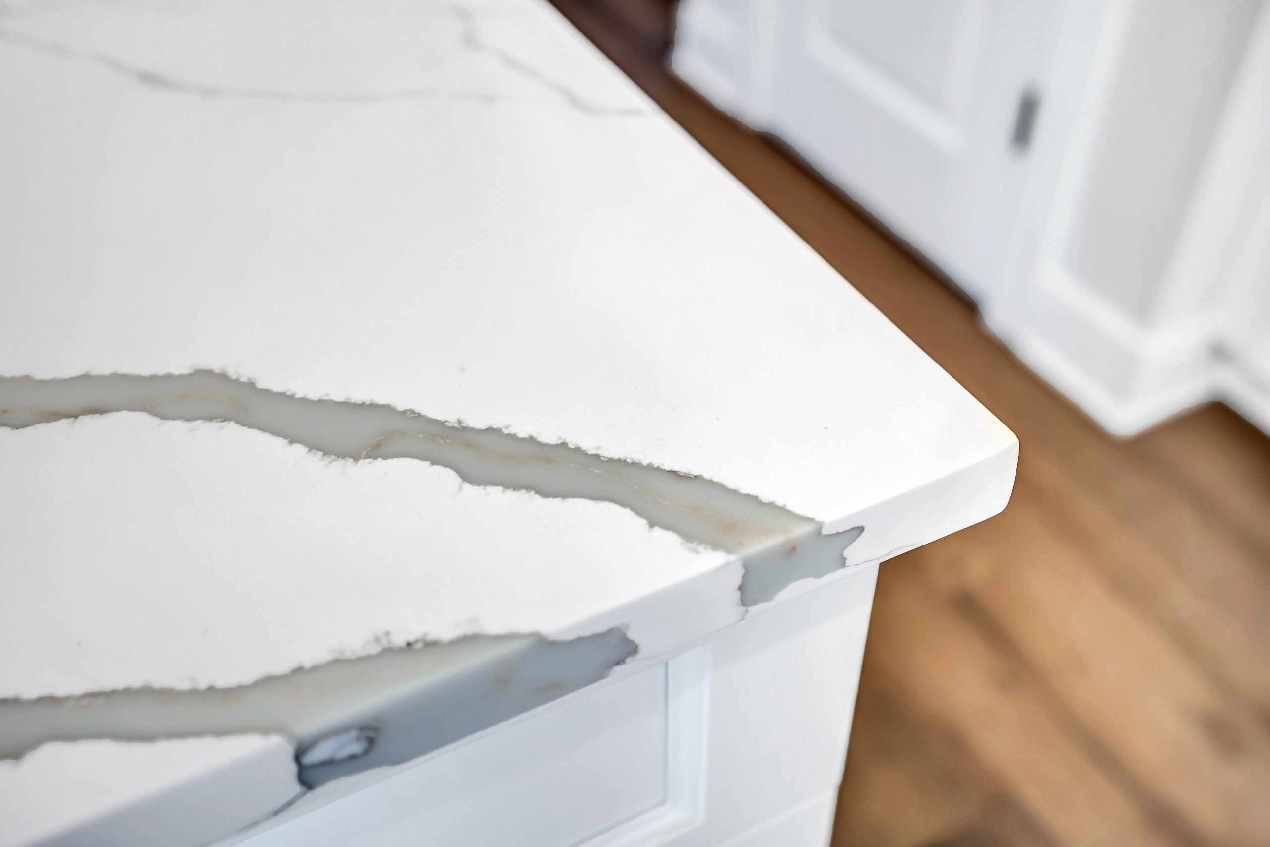 Close-up of a white marble countertop with subtle gray veining, featuring a smooth, polished surface and a clean, sharp edge. The background shows a blurred wooden floor and white cabinetry.