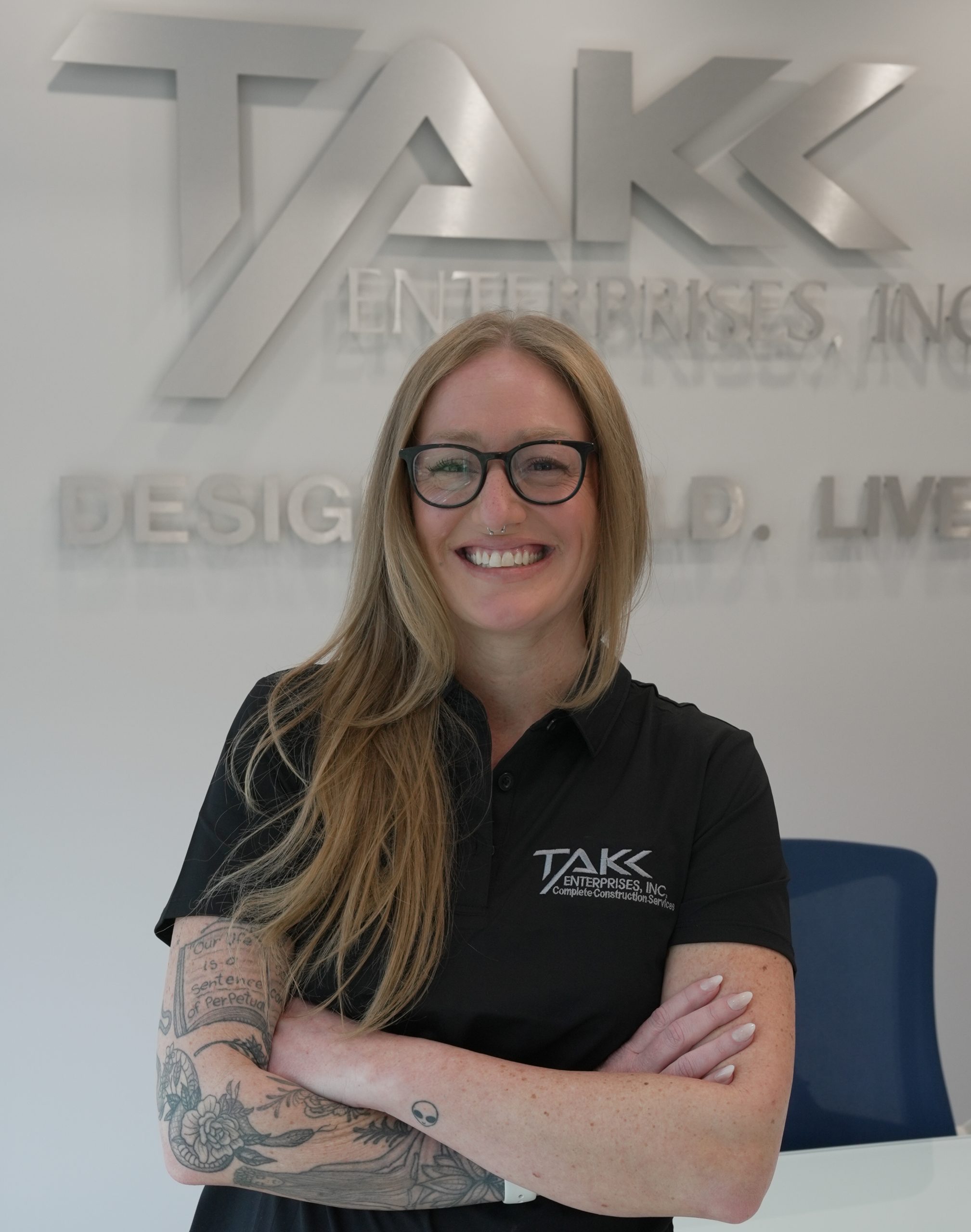 DSC00168 A woman with long blonde hair and glasses smiles with arms crossed, standing in front of a TAKK Enterprises, Inc. sign. She wears a black polo shirt and has tattoos on her left forearm.
