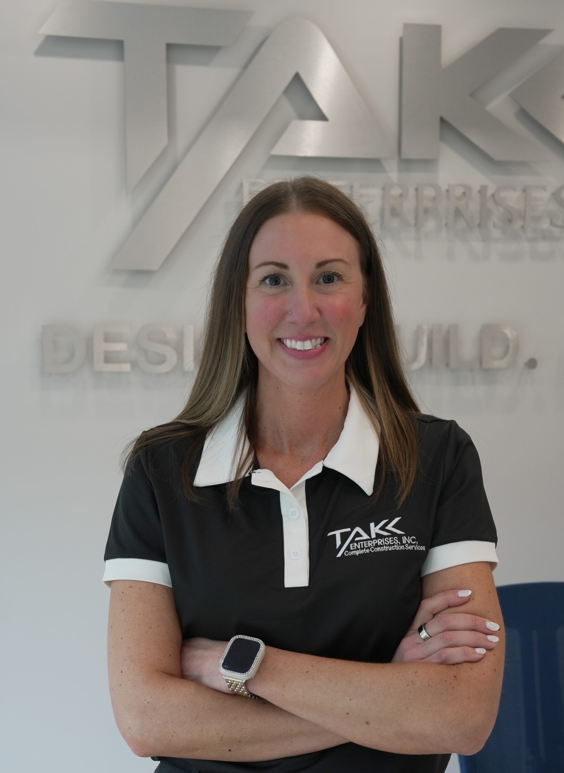 DSC00175 A woman with long brown hair stands with her arms crossed, smiling. She wears a black and white collared shirt with a TAKK Enterprises, Inc. logo and a smartwatch. A silver TAKK Enterprises sign is on the wall behind her.