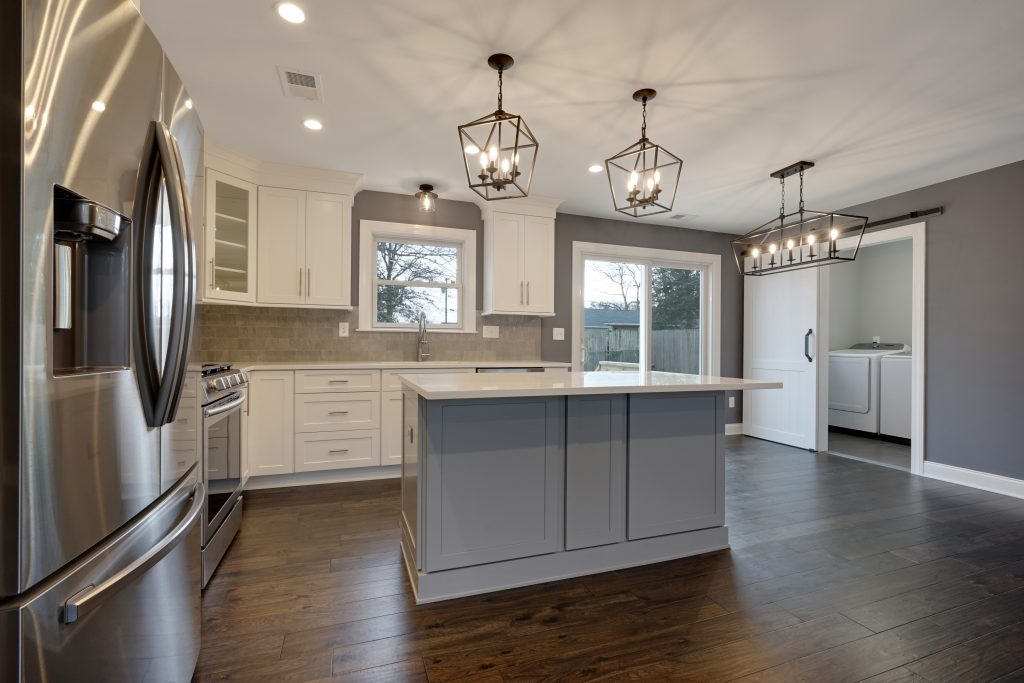 Modern kitchen with white cabinets, a gray island, stainless steel appliances, pendant lights, wooden floors, and an open view into a laundry room with washer and dryer. Large windows let in natural light.