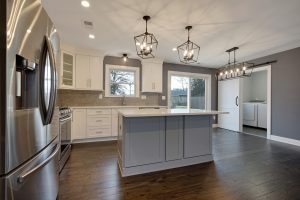 Modern kitchen with white cabinets, a gray island, stainless steel appliances, pendant lights, wooden floors, and an open view into a laundry room with washer and dryer. Large windows let in natural light.