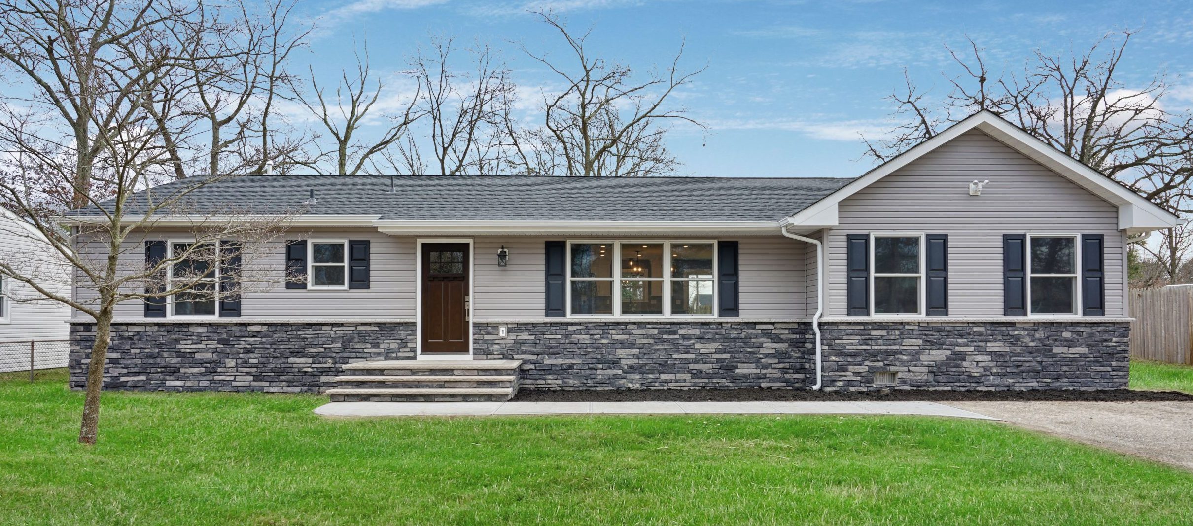 Single-story house with gray siding and stone facade, black shutters, a dark brown front door, and steps leading to the entrance. The yard has green grass and leafless trees, with a driveway on the right.