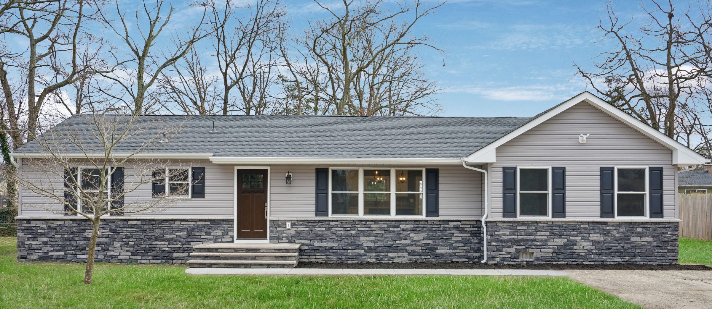 Single-story house with gray siding and dark stone accents, black shutters, and a brown front door. The house has a front lawn, a bare tree, and leafless trees in the background under a blue sky.