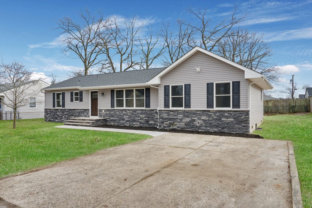 Single-story suburban house with gray siding and stone accents, large front windows, and a concrete driveway. The lawn is green and trees without leaves are in the background under a blue sky.