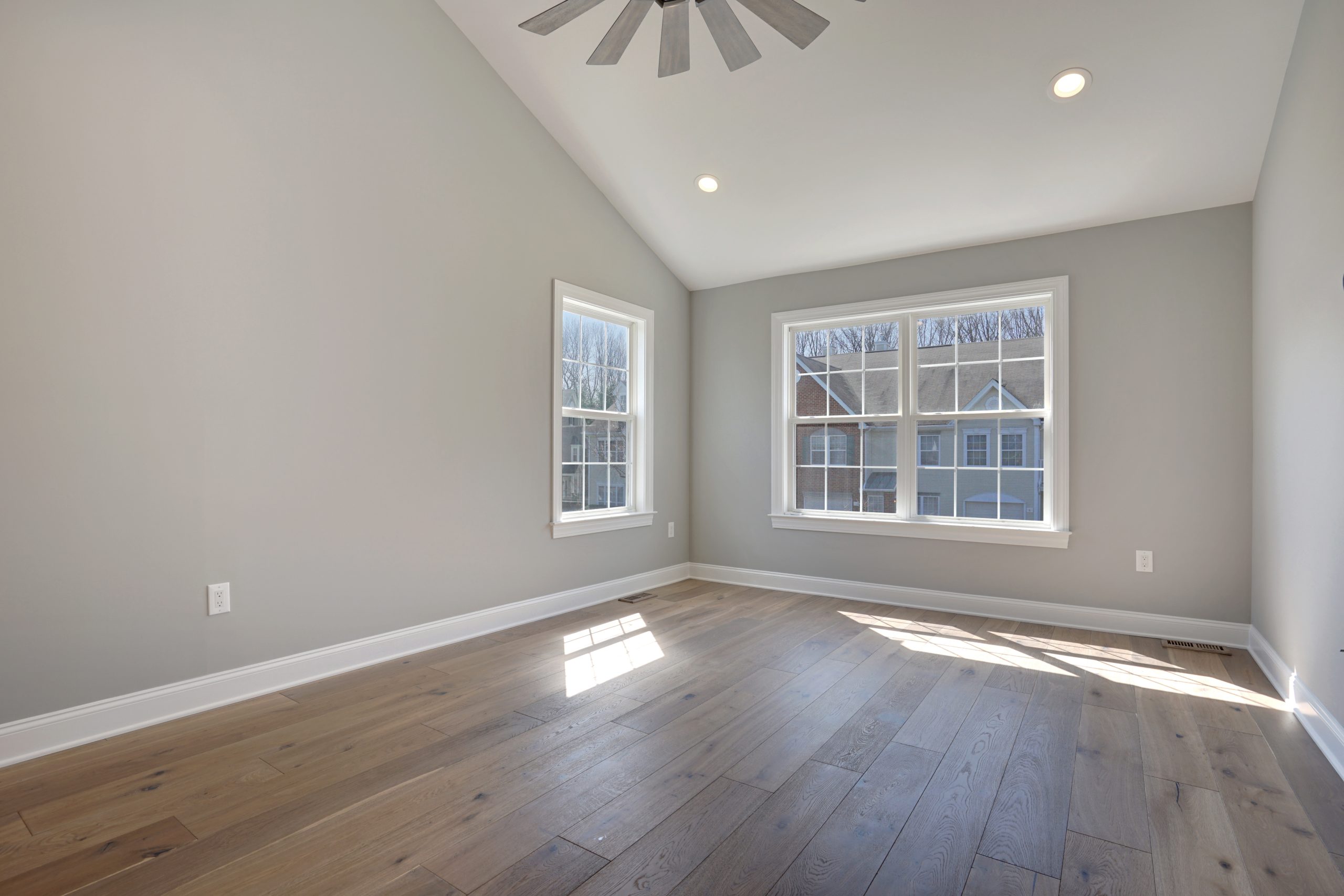 Bright, empty room with light gray walls, wide plank wood flooring, large double windows letting in sunlight, and a ceiling fan with recessed lighting on a vaulted ceiling.