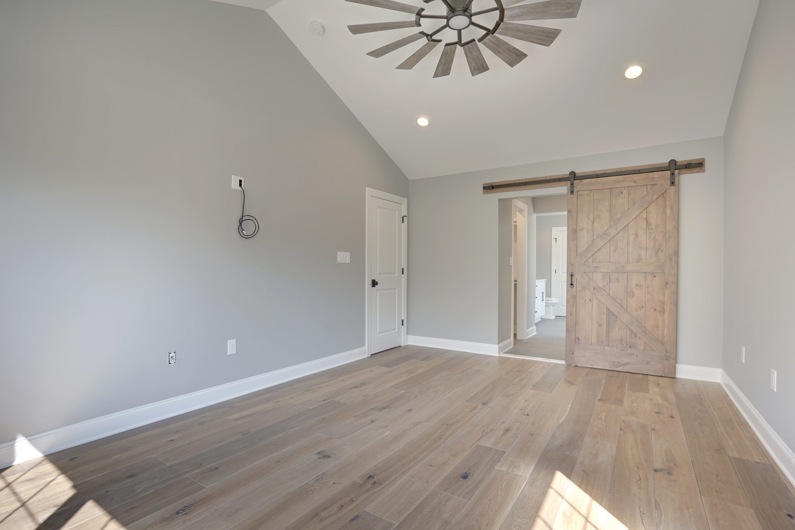 A modern empty room with light wood flooring, gray walls, a ceiling fan, recessed lighting, and a rustic wooden sliding barn door leading to another room. Sunlight streams in through a window out of view.