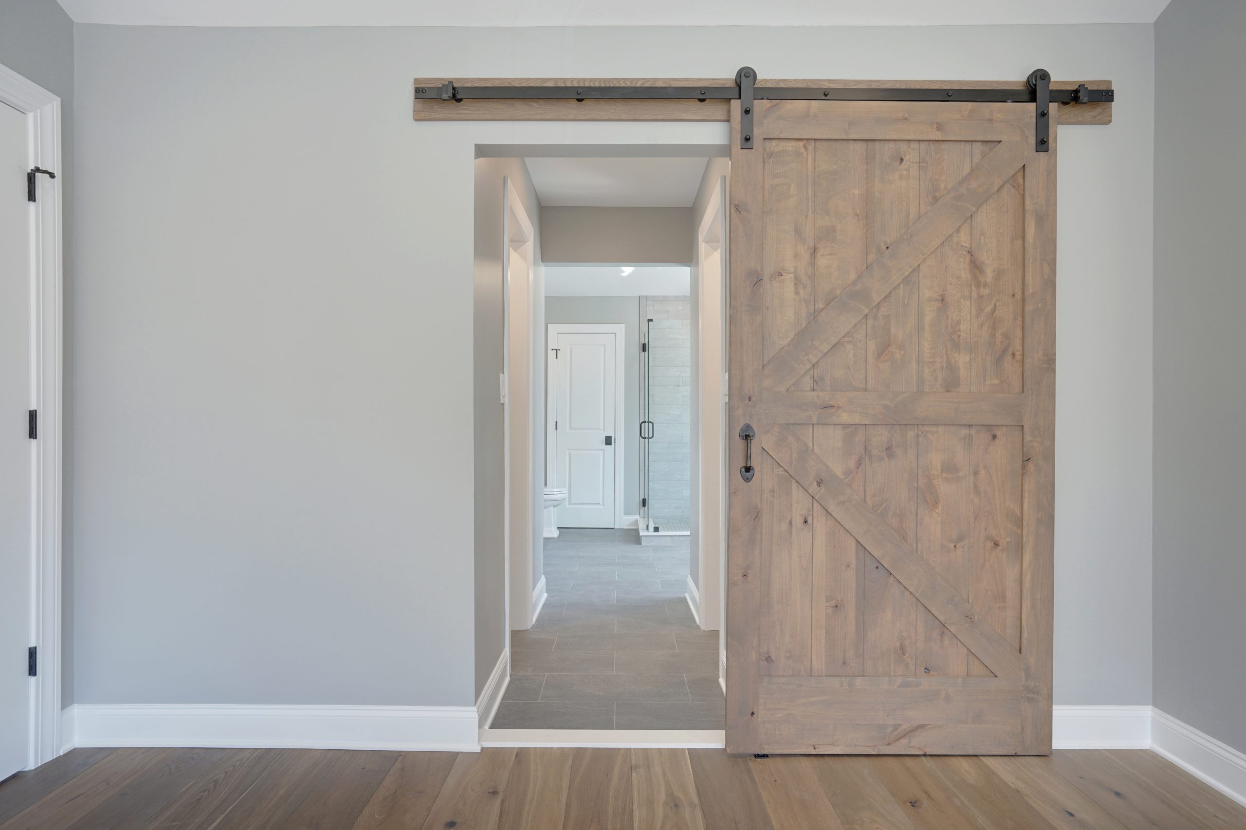 A wooden sliding barn door partially open, revealing a hallway with light gray walls and tile flooring, leading to a white door and a glass shower door in the background.
