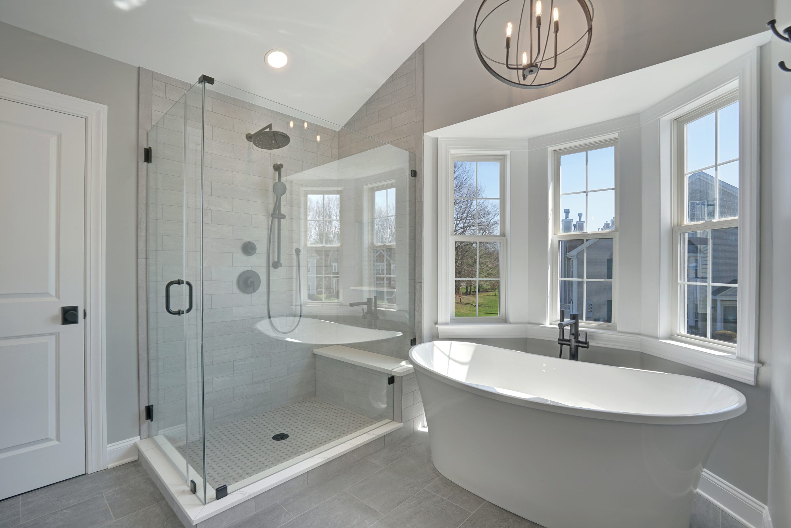 Modern bathroom with a glass-enclosed shower, a freestanding white bathtub by large bay windows, gray tiled floor, and a round chandelier hanging from the ceiling. Natural light fills the space.