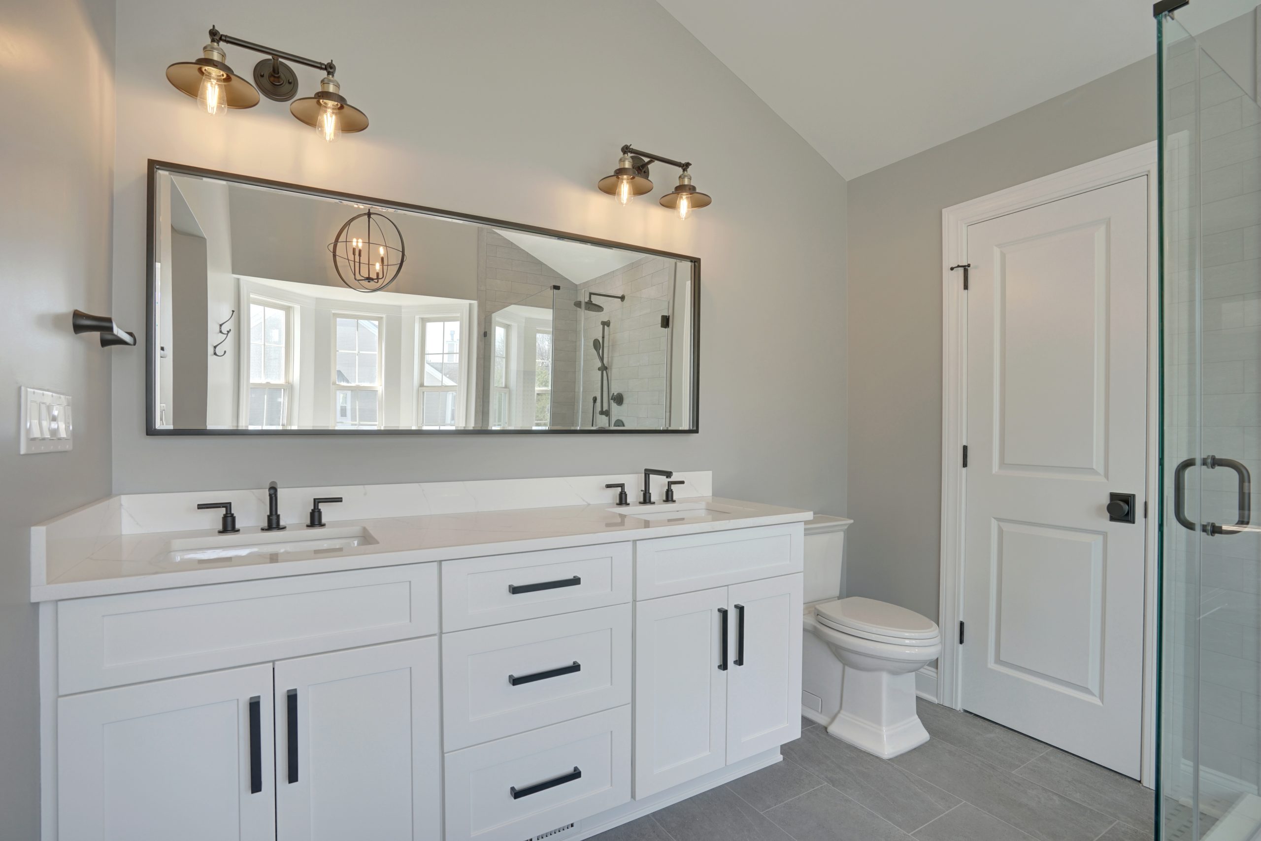 Modern bathroom with a double-sink white vanity, large rectangular mirror, two black light fixtures, a white toilet, gray tile floor, glass shower door, and gray walls. Natural light comes from windows in the background.