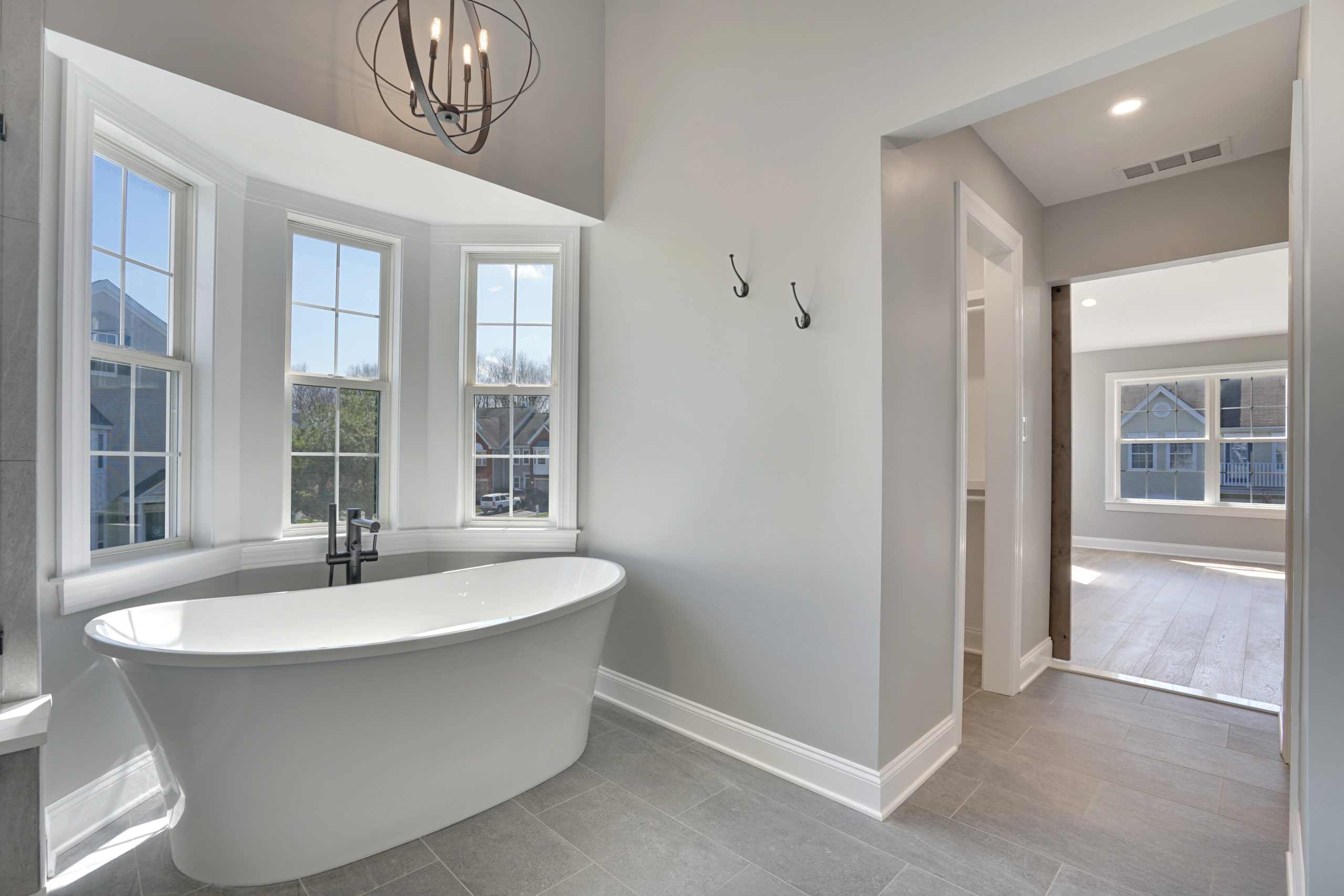 Modern bathroom with a freestanding white bathtub by large bay windows, gray tile floor, light gray walls, two wall hooks, and an open doorway leading to a bright room with wood flooring.
