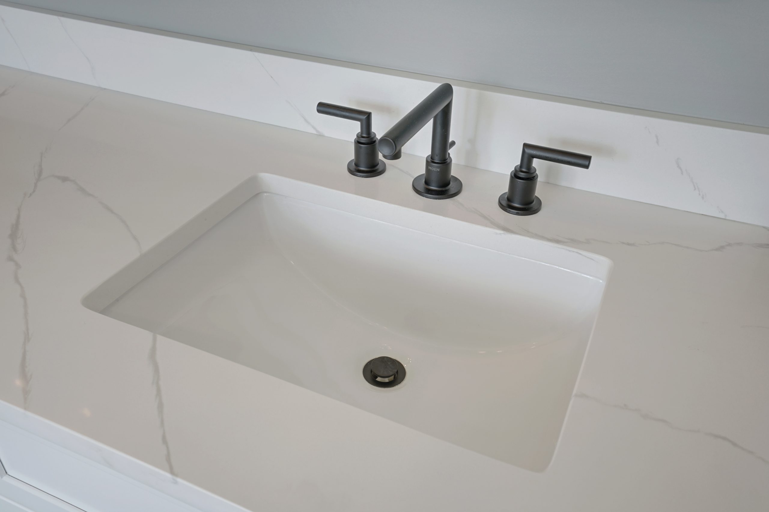A modern white bathroom sink with a rectangular basin, black faucet, and two matching handles set in a marble countertop with subtle gray veining.