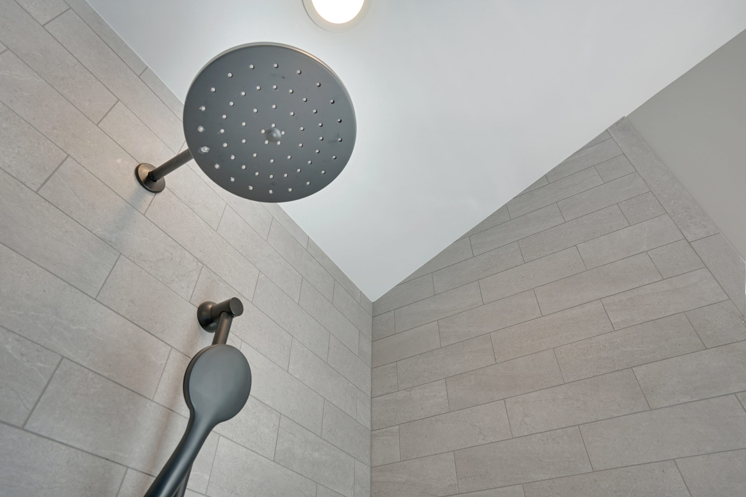 Modern shower with a large round rain showerhead and handheld attachment, both mounted on a tiled wall with light gray rectangular tiles. A ceiling light is visible above the showerhead.