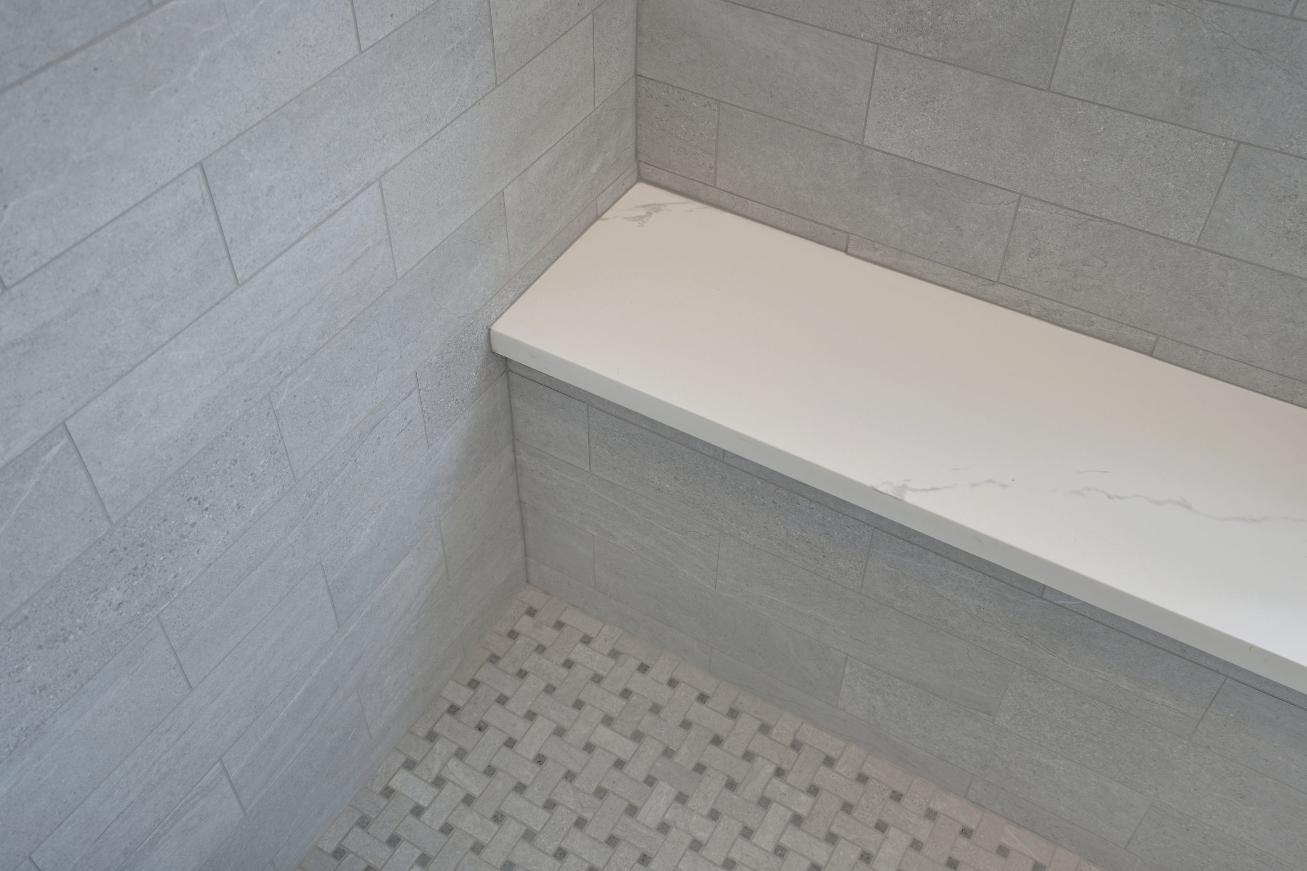 A shower corner with gray rectangular wall tiles, a built-in bench with a white marble top, and a floor featuring a woven pattern of small square tiles in light and dark gray tones.