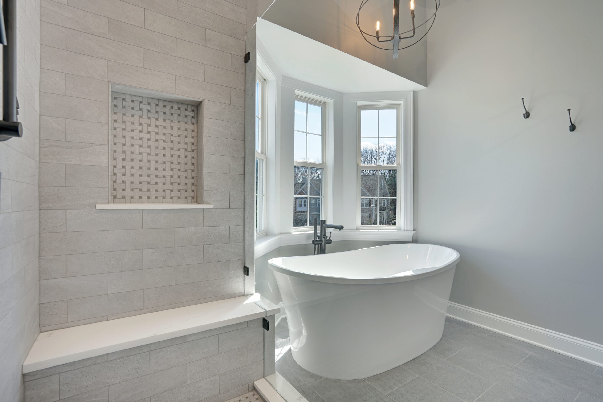 Modern bathroom with a white freestanding bathtub by large windows, light gray tiled walls, a built-in shower bench, and a decorative tile niche. Natural light fills the space.