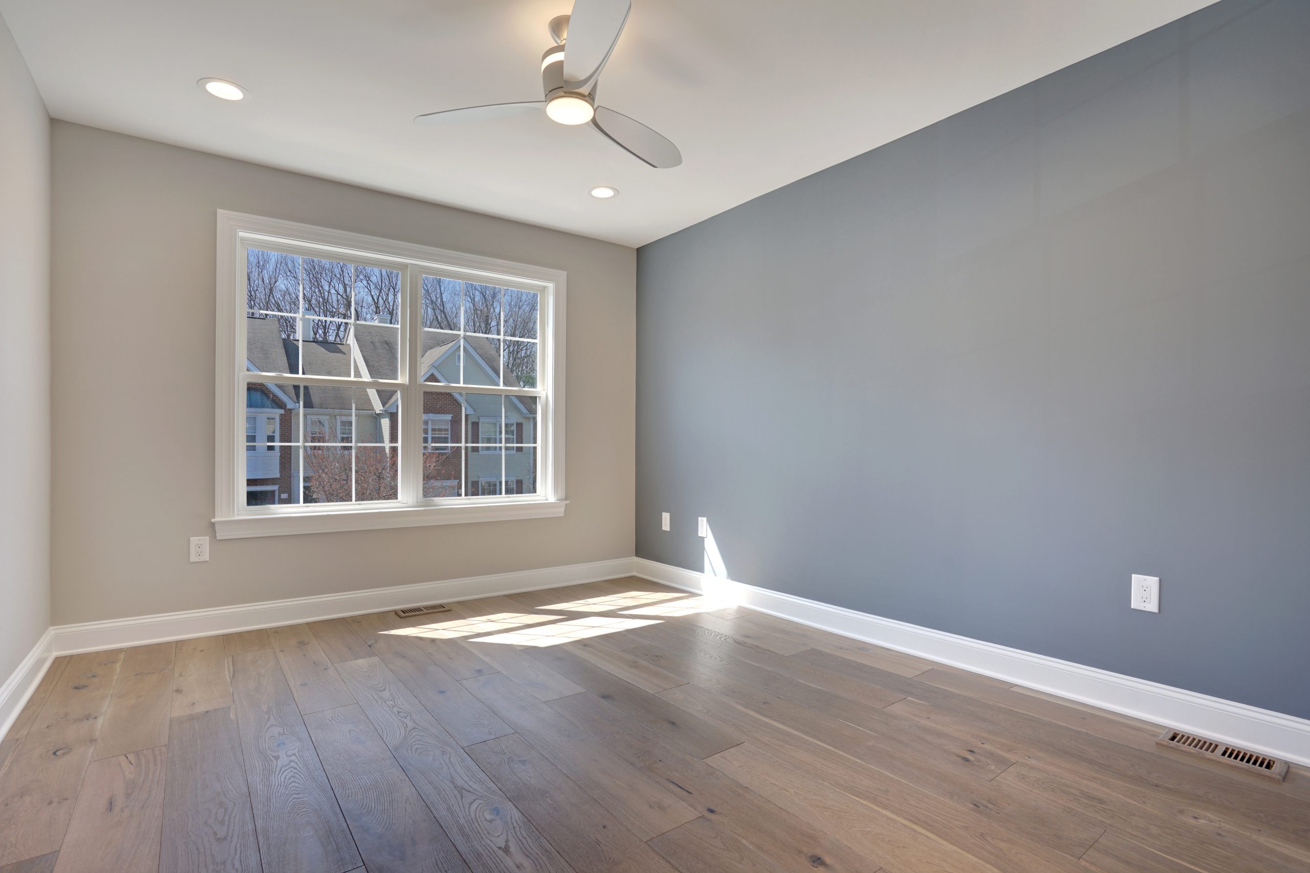 Empty, modern room with light wood floors, gray accent wall, white trim, a ceiling fan with a light, and a large window letting in natural sunlight. Some houses and trees are visible outside the window.