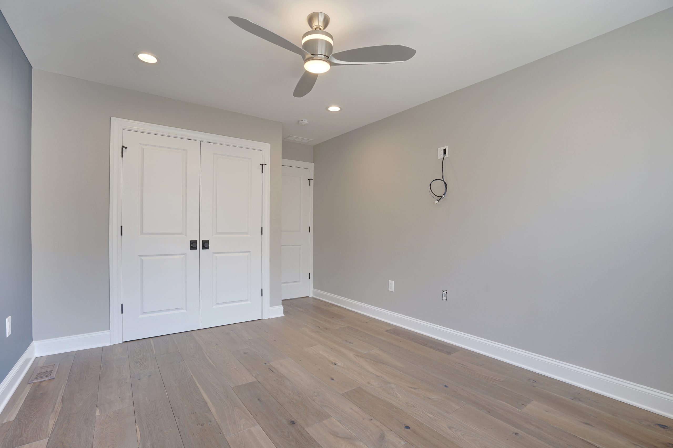 Empty modern room with light gray walls, wood floor, double white closet doors, a ceiling fan with lights, and electrical outlets on the wall. A wire hangs near the center, possibly for a TV or device.