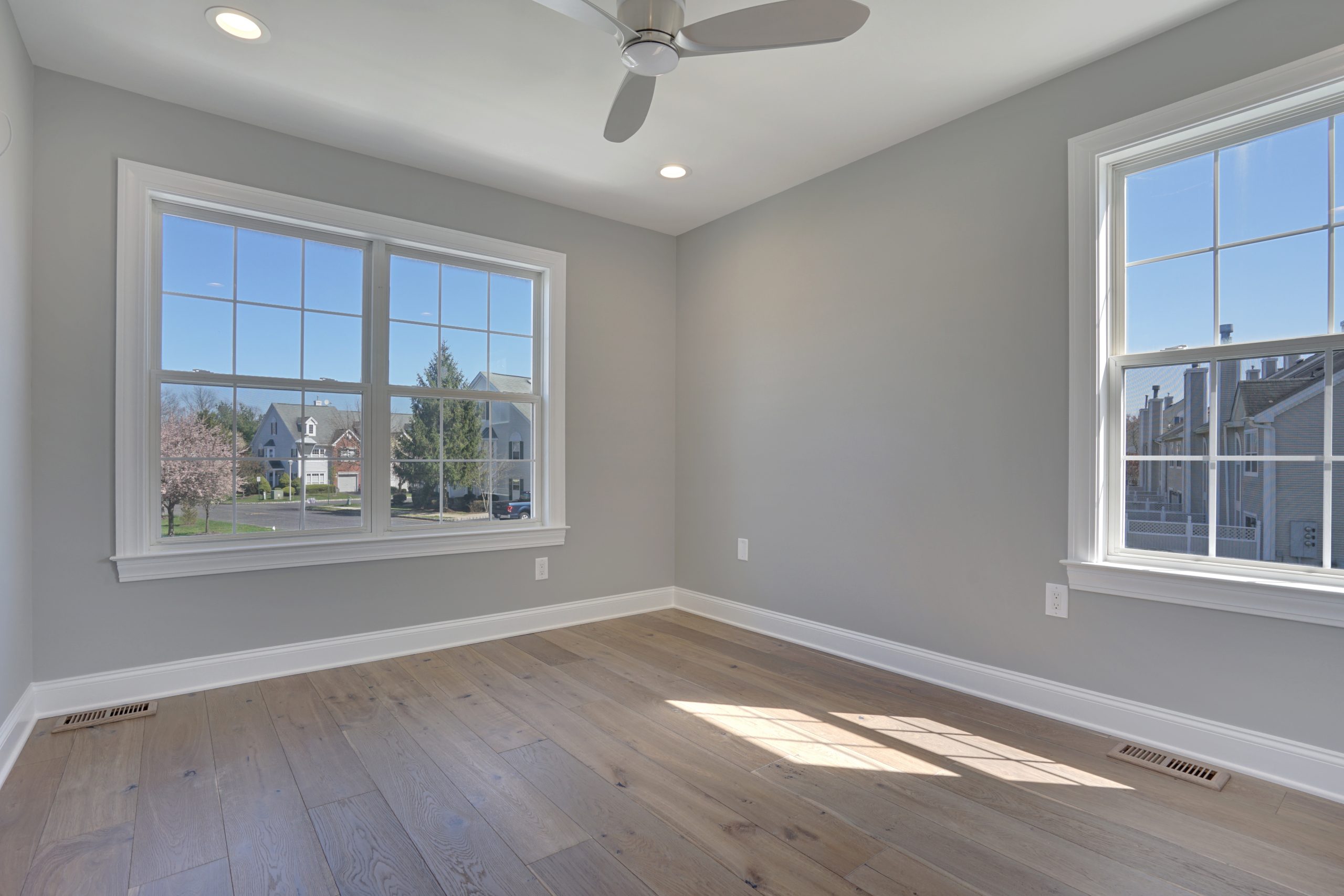 Bright, empty room with light gray walls, wood floors, two large windows letting in natural light, ceiling fan, and recessed lighting; view of houses and trees outside.