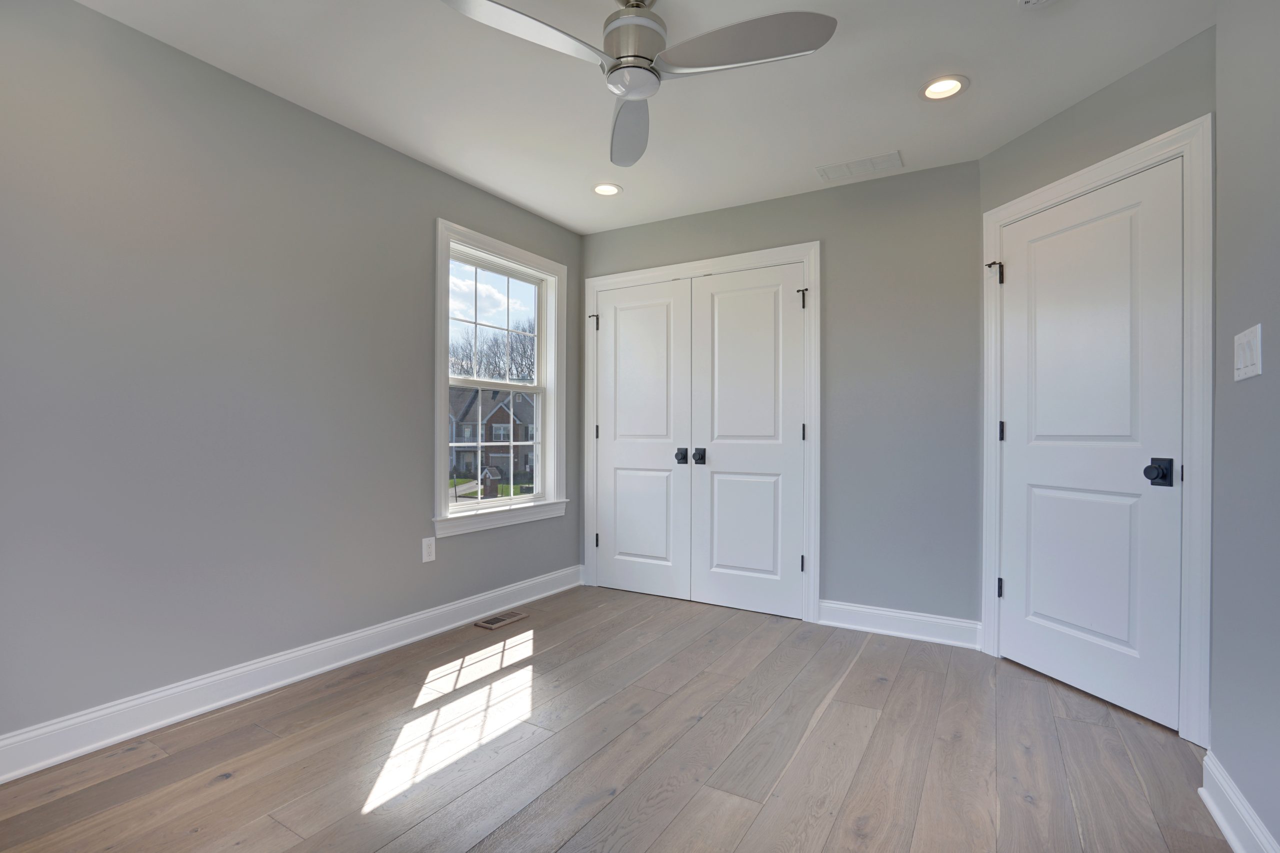 A bright, empty room with light gray walls, light wood flooring, a large window letting in sunlight, a ceiling fan, and two white doors with black handles.