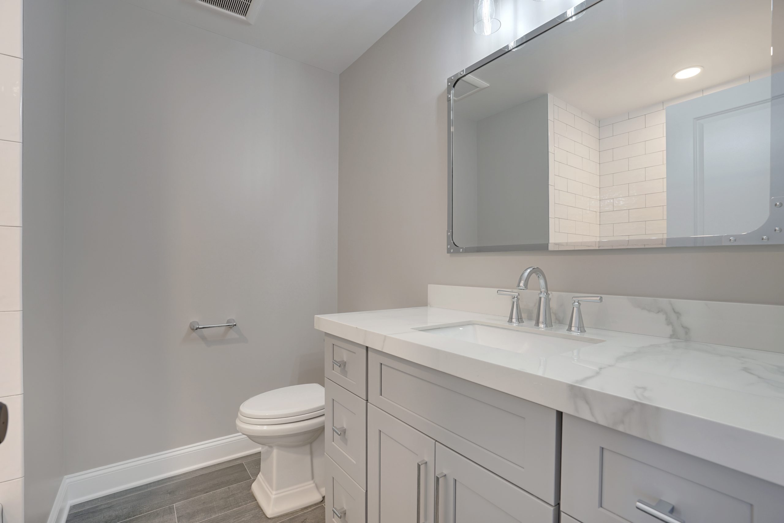 Modern bathroom with light gray walls, a white vanity with a marble countertop, chrome faucet, rectangular mirror, white toilet, and gray tile flooring. The room has recessed lighting and a simple, clean design.