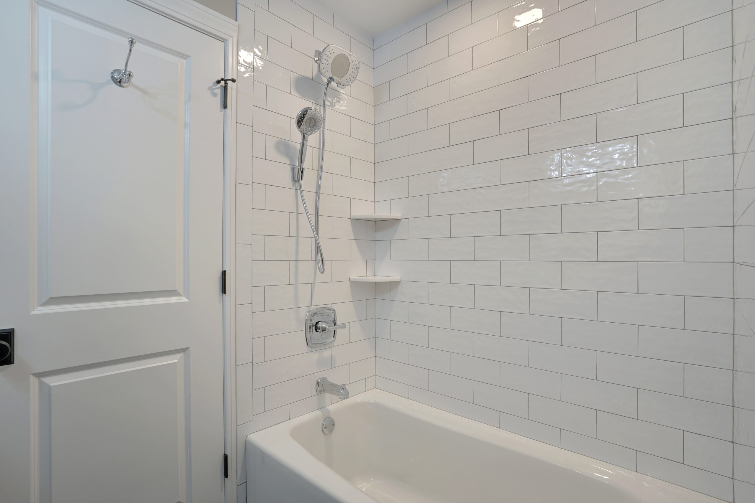 A white bathroom with a rectangular bathtub, chrome showerhead and faucet, white subway tile walls, two built-in corner shelves, and a white door with a towel hook.