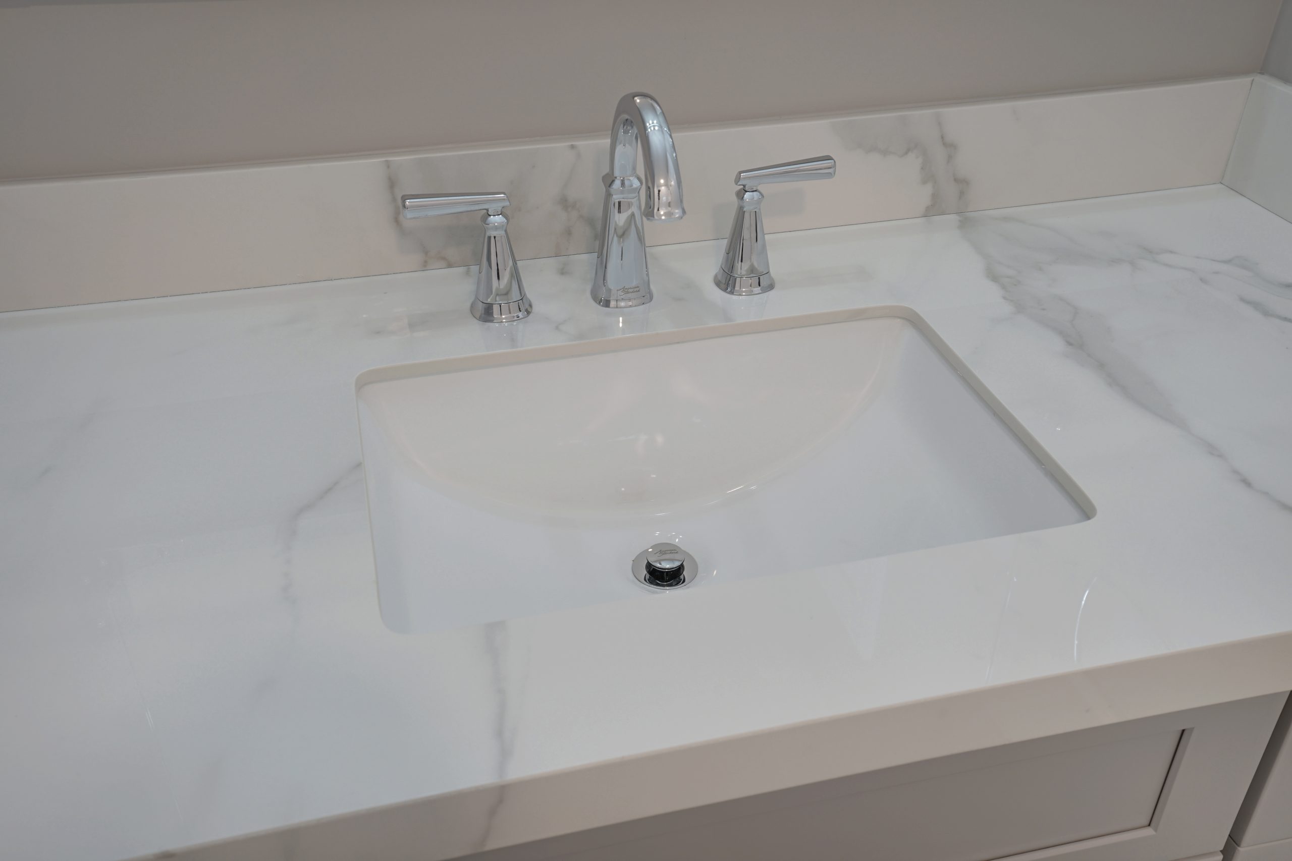 A modern, rectangular white sink with a chrome faucet and handles is set in a glossy white marble countertop with grey veining. The backsplash and surrounding counter are also made of matching marble.