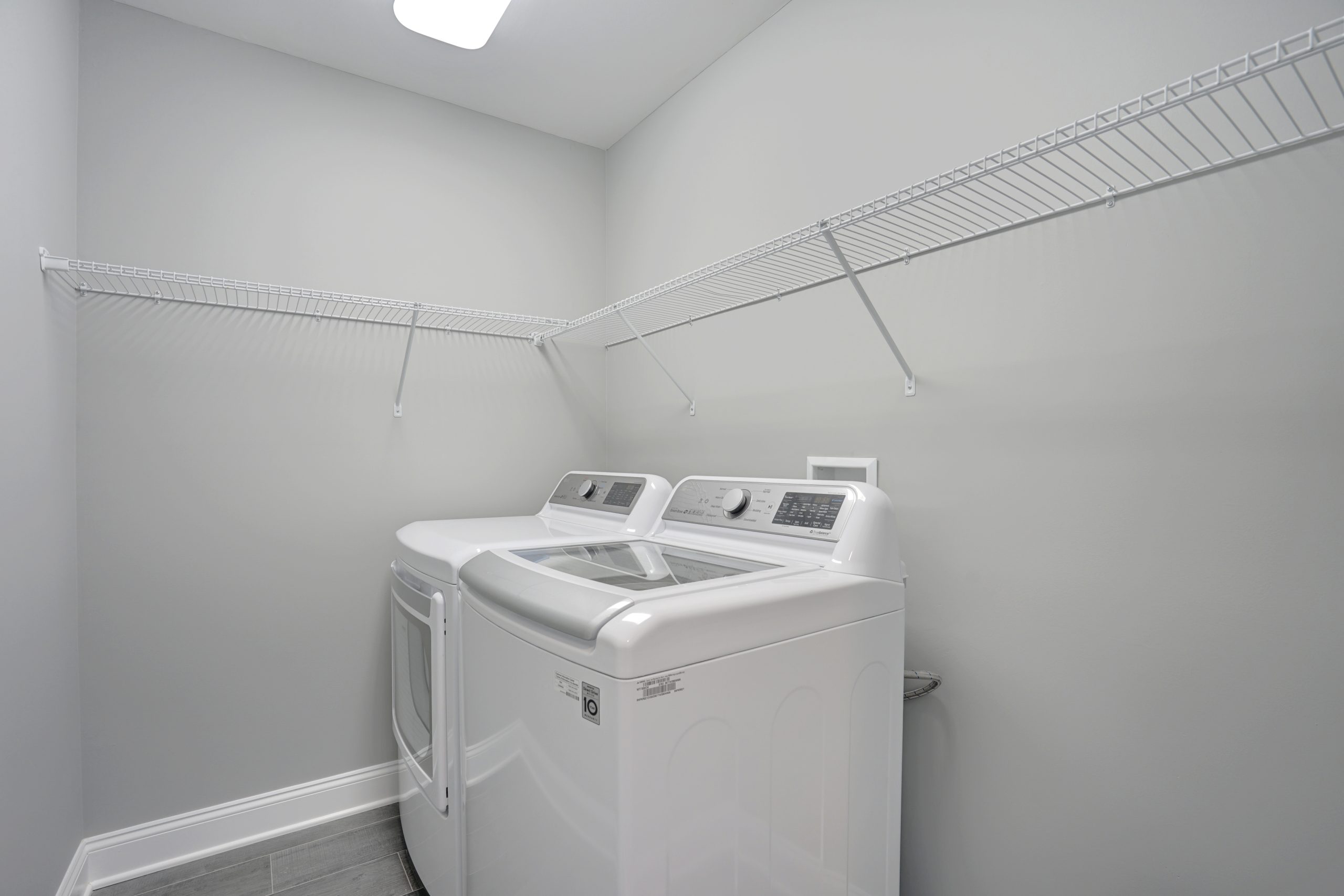 A modern laundry room with a white washing machine and dryer side by side, light gray walls, tiled floor, and two white wire shelves mounted on the walls above the appliances.