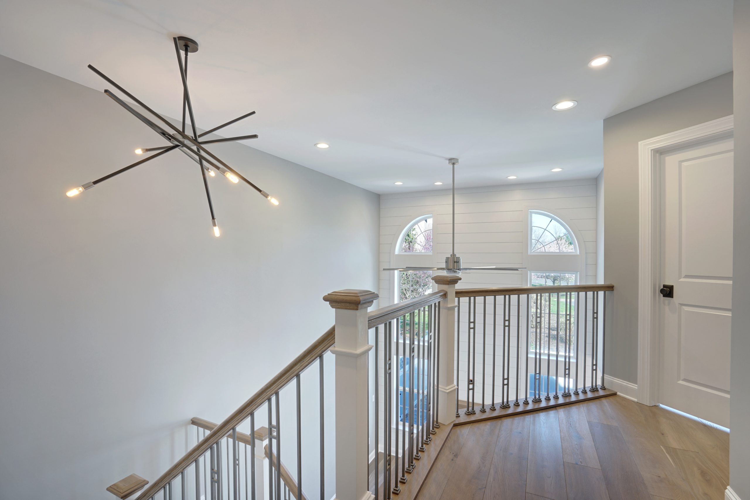 A modern staircase landing with wood floors, metal railings, a large geometric light fixture, and arched windows letting in natural light. Walls are light gray and white, with recessed ceiling lights.