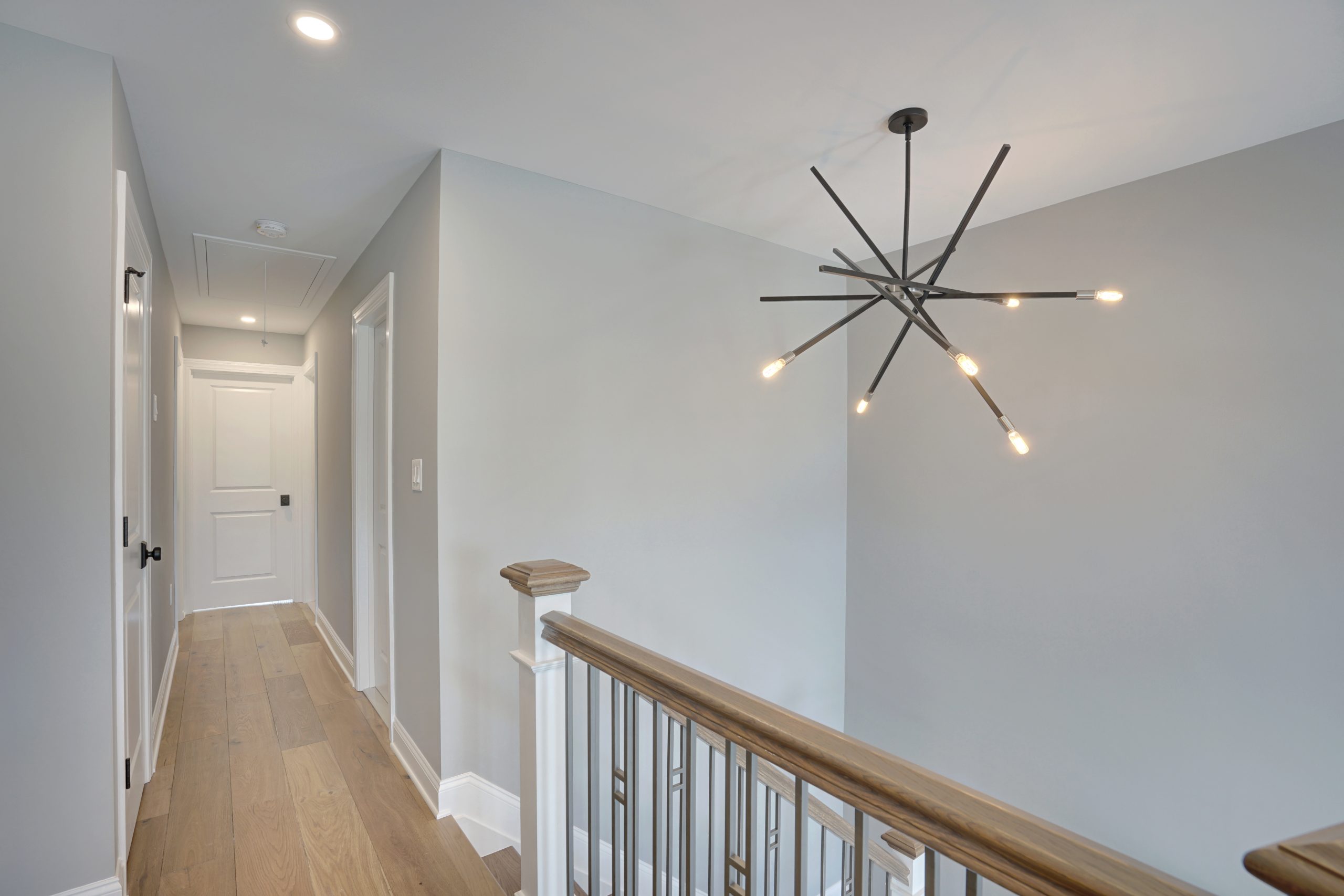 A modern hallway with light gray walls, light wood flooring, white doors, and a contemporary black chandelier with exposed bulbs hanging above a staircase with a wooden handrail.