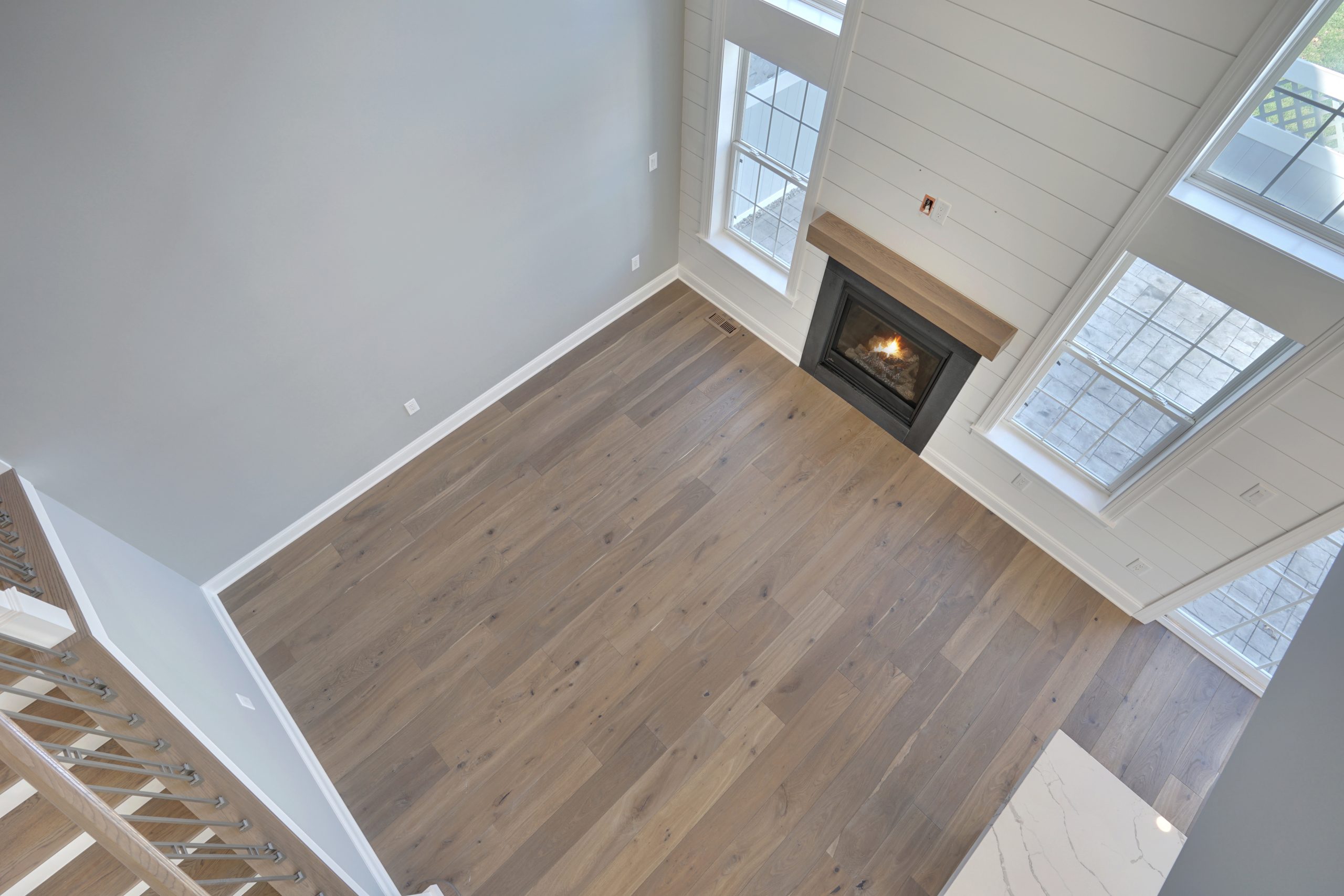 A bright, modern living room with light wood flooring, large windows, a white shiplap wall, and a fireplace with a wooden mantel, viewed from above near a staircase.