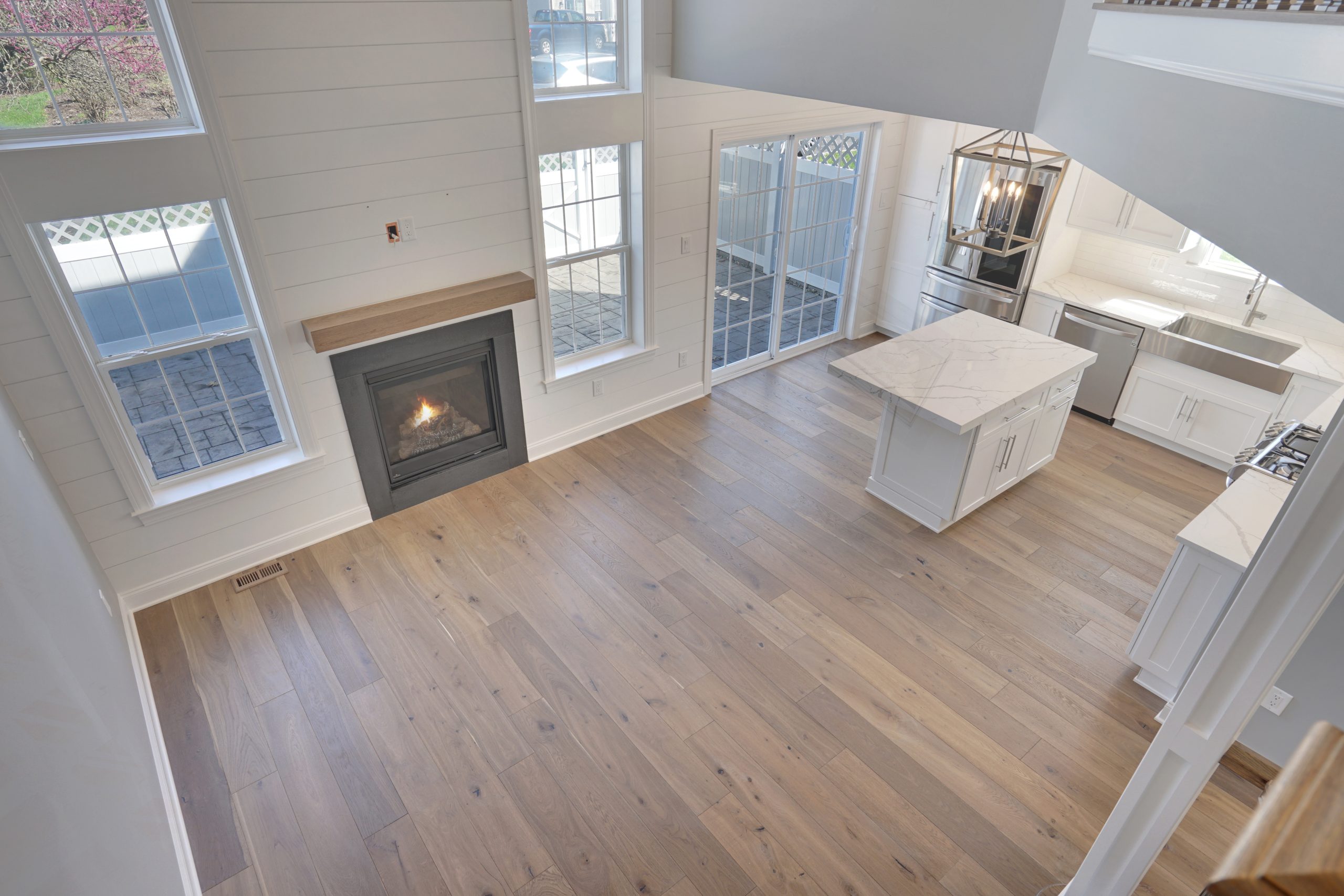 Spacious, modern open-concept kitchen and living area with light wood floors, a fireplace, large windows, an island with a marble countertop, and stainless steel appliances.