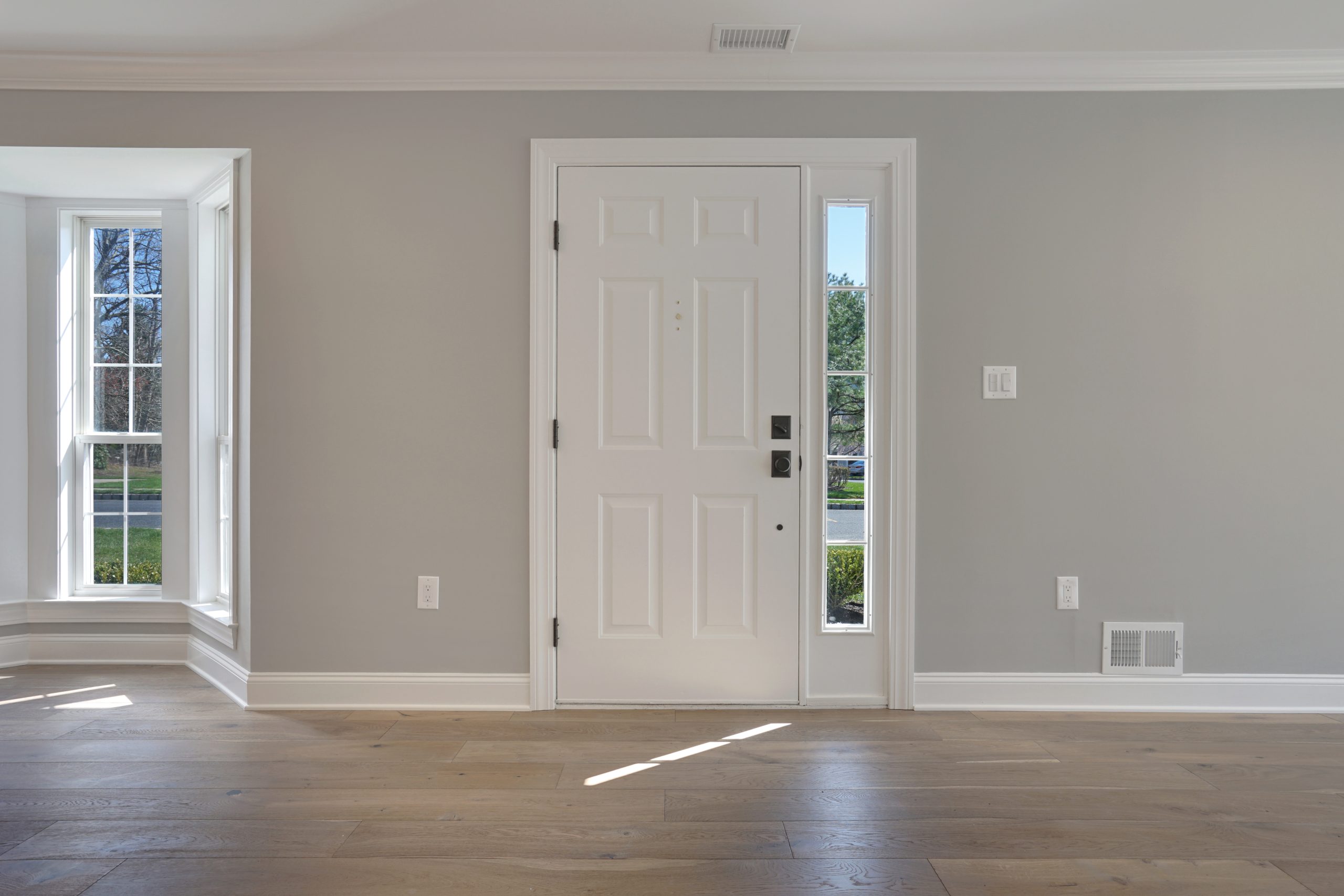 A bright, empty room with light wood floors, gray walls, and a white front door with glass side panels. Sunlight streams in through a large bay window on the left.
