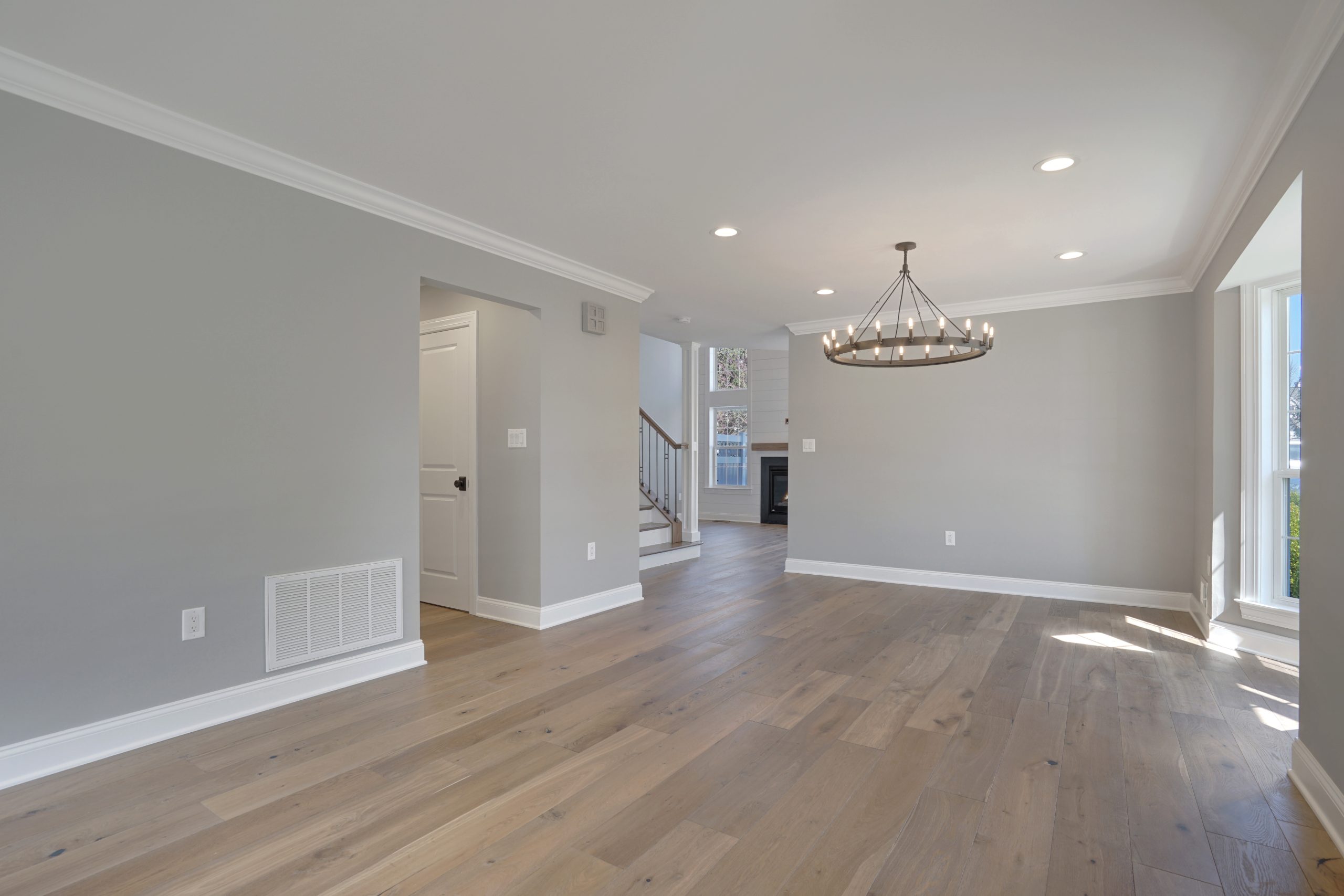 Bright, empty living room with light gray walls, large windows, wood flooring, recessed lighting, and a modern chandelier. The space opens into a hallway with stairs and a visible fireplace in the background.