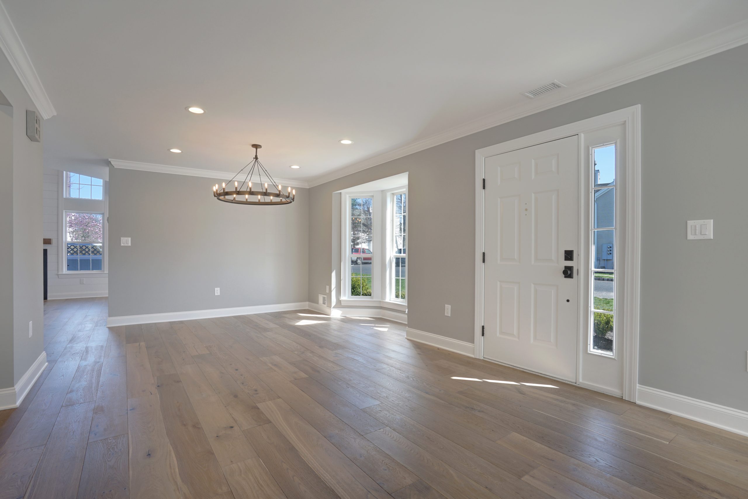 A bright, empty room with light wood floors, gray walls, white trim, a large bay window, a white front door, and a modern circular chandelier hanging from the ceiling. Sunlight streams through the windows.