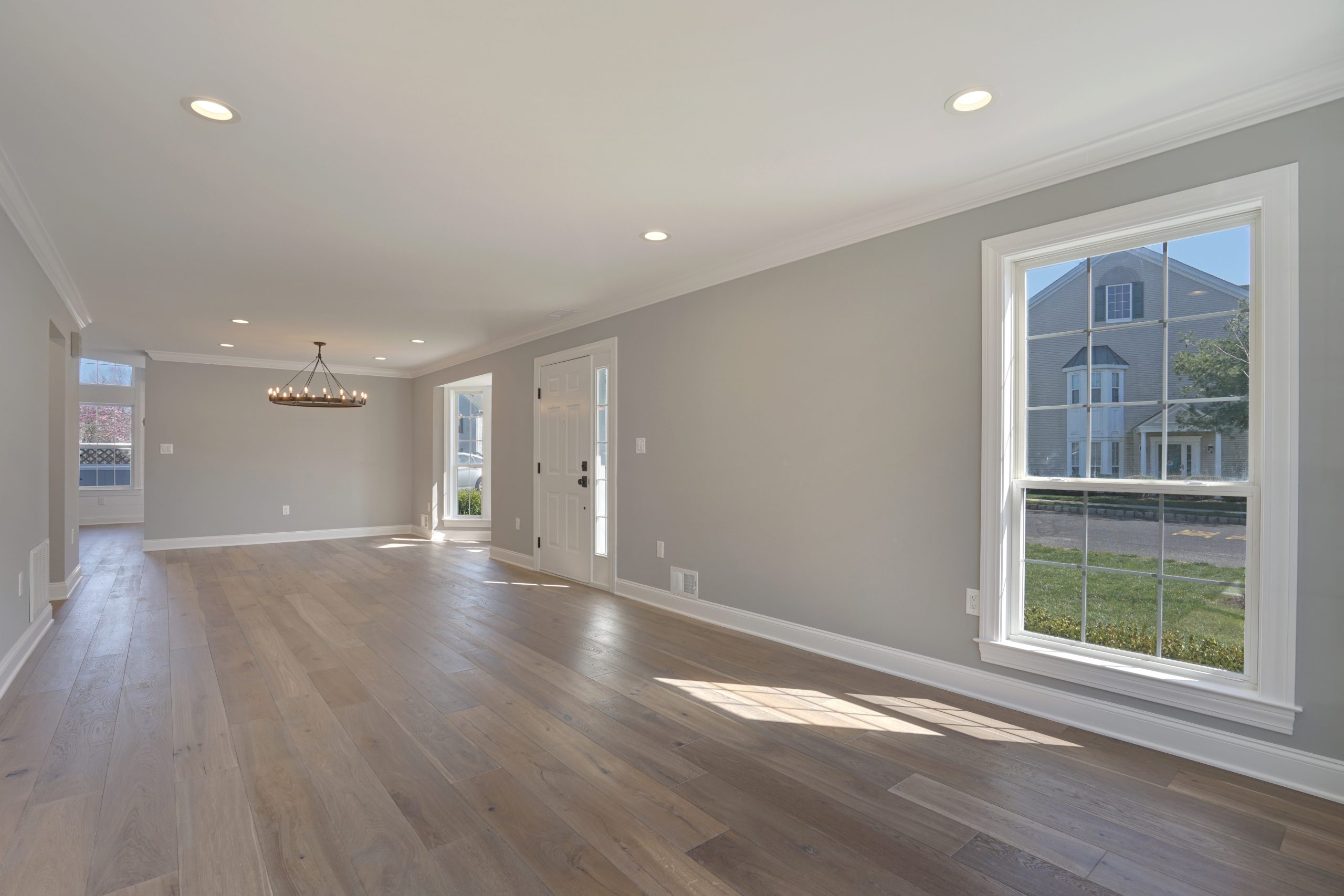 A spacious, empty living room with light wood floors, gray walls, recessed lighting, a modern chandelier, large windows letting in natural light, and a white exterior door. A neighboring house is visible through the windows.