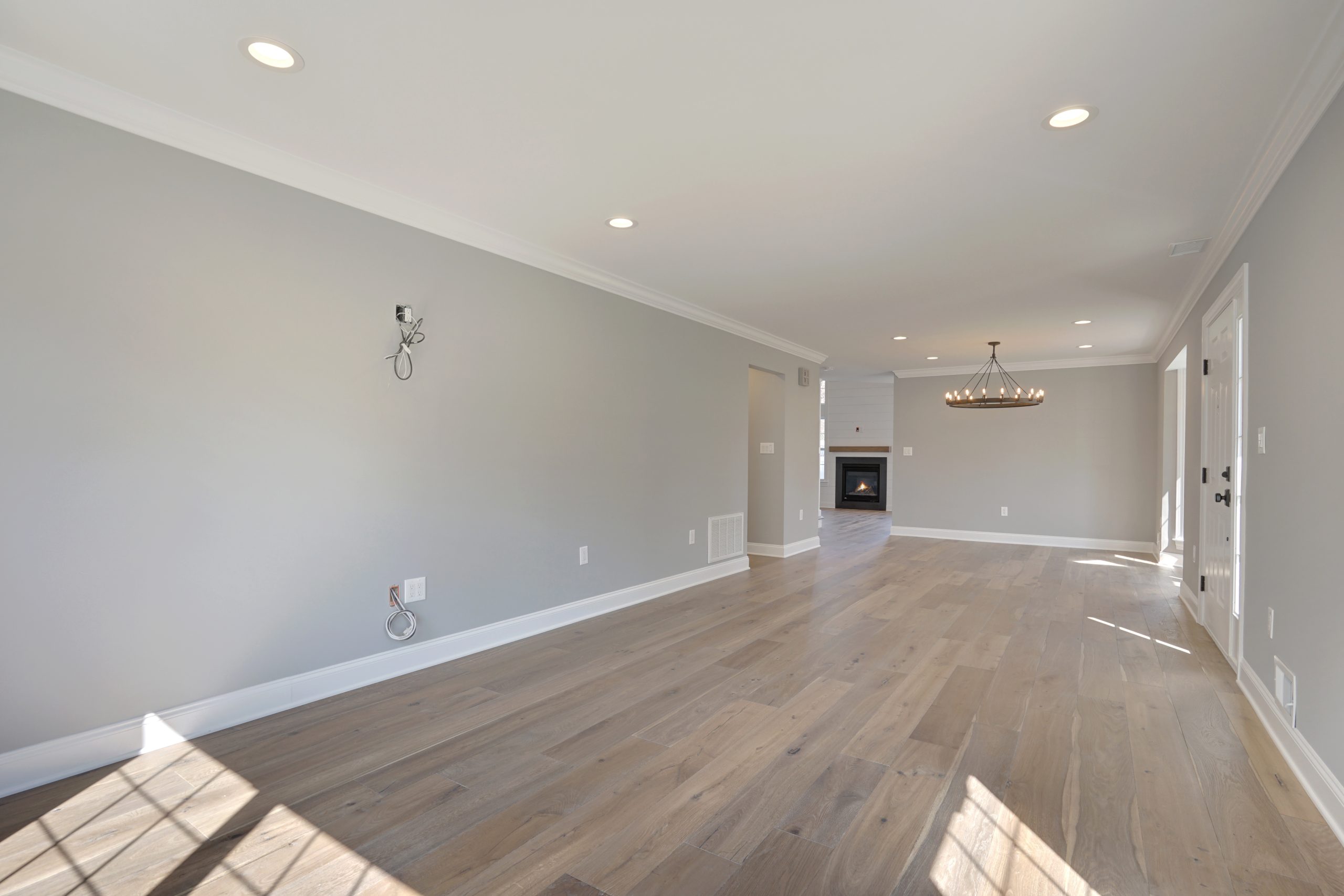 Spacious, empty living room with light wood flooring, light gray walls, recessed lighting, and large windows letting in natural light. Fireplace and chandelier are visible in the background.