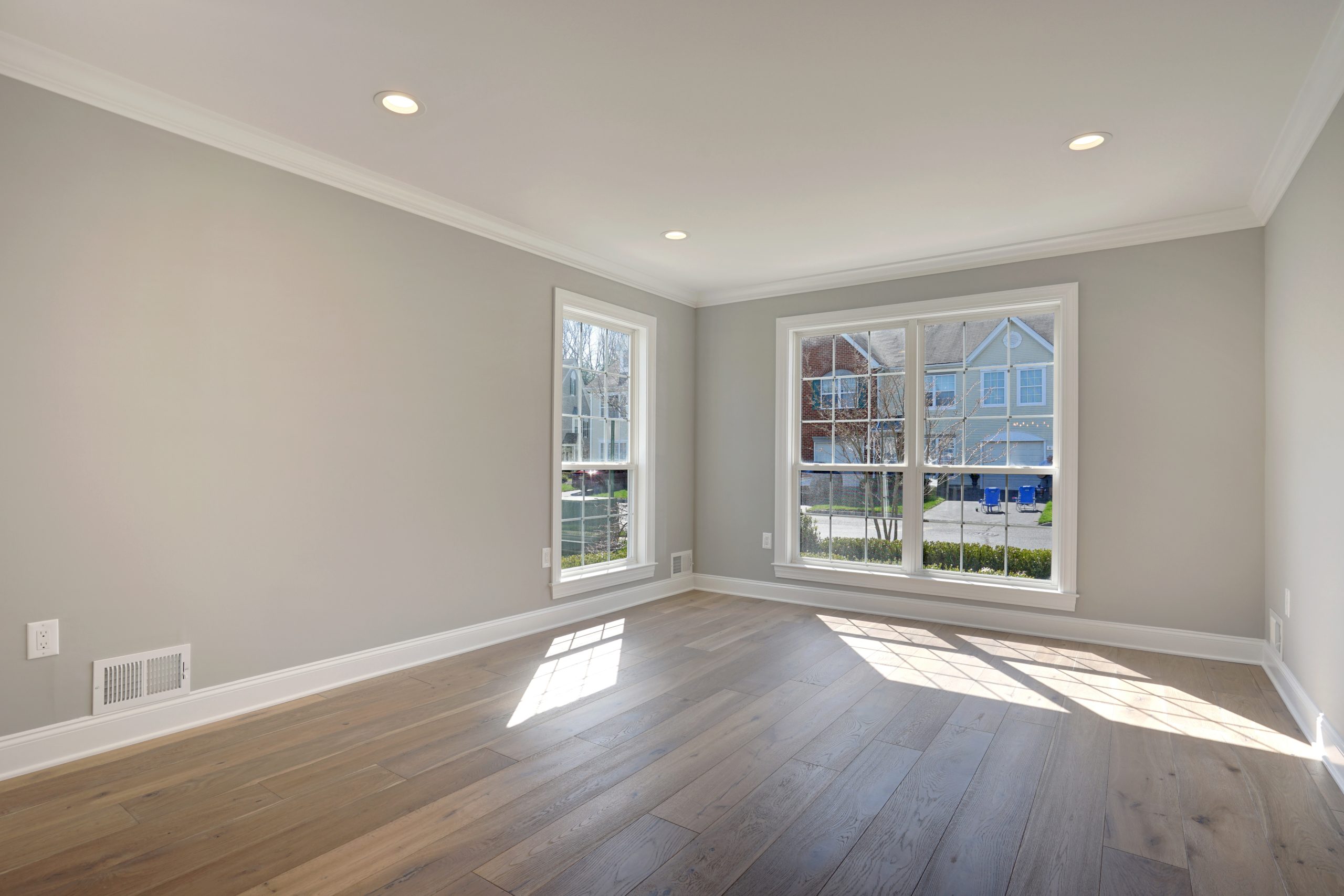 Bright, empty room with light gray walls, recessed ceiling lights, large windows, and light wood flooring. Sunlight streams in, and neighboring houses are visible outside.