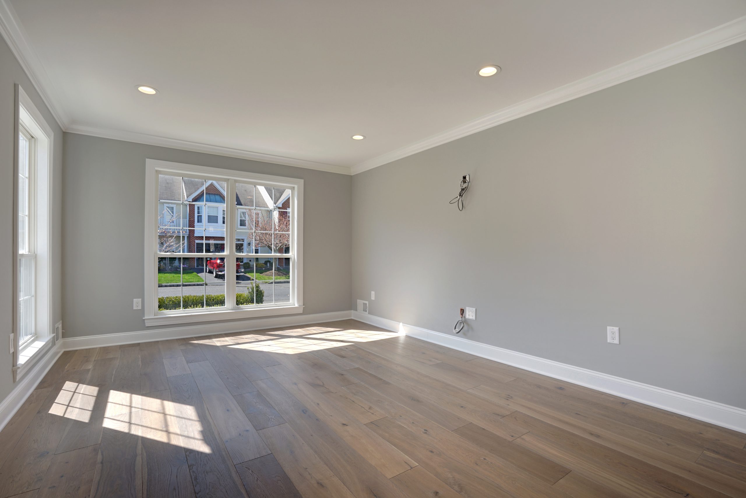 Empty living room with light gray walls, wide windows letting in natural light, recessed ceiling lights, and light wood flooring. The windows provide a view of houses and greenery outside.