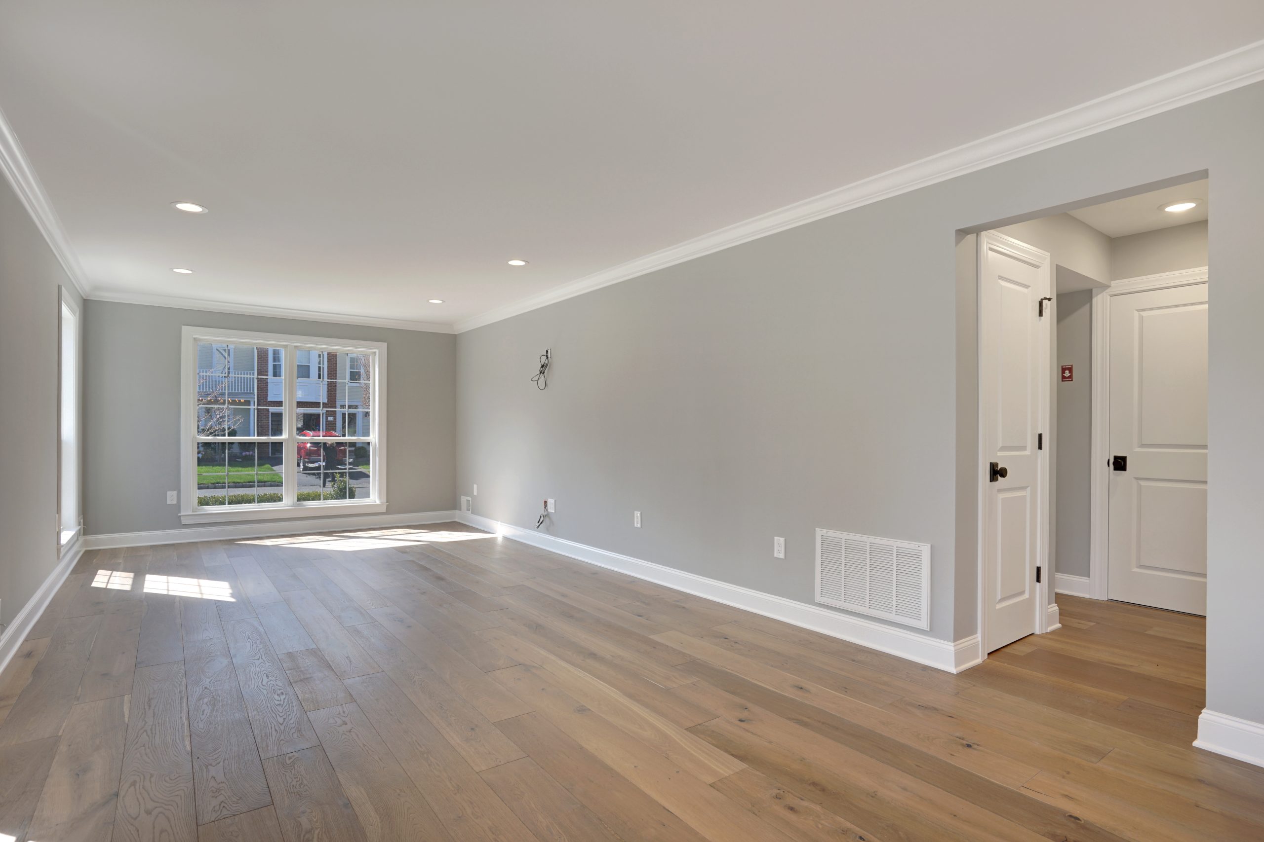 Spacious, empty living room with light gray walls, recessed lighting, large window letting in natural light, and light wood flooring. There are white baseboards, crown molding, and an open doorway leading to a hallway.
