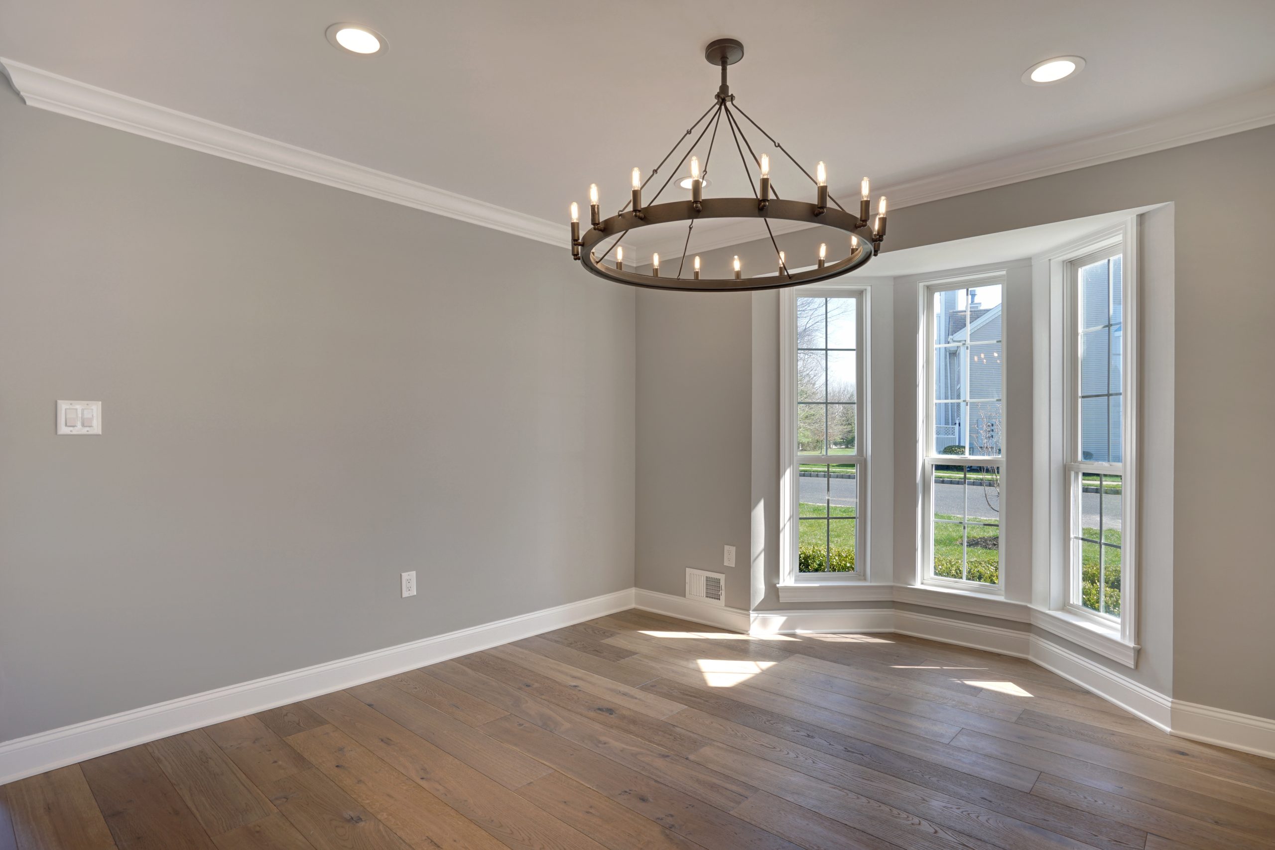 Bright, empty room with light gray walls, large bay window, wooden floor, and circular black chandelier with candle-style lights hanging from the ceiling. Sunlight streams through the window onto the floor.