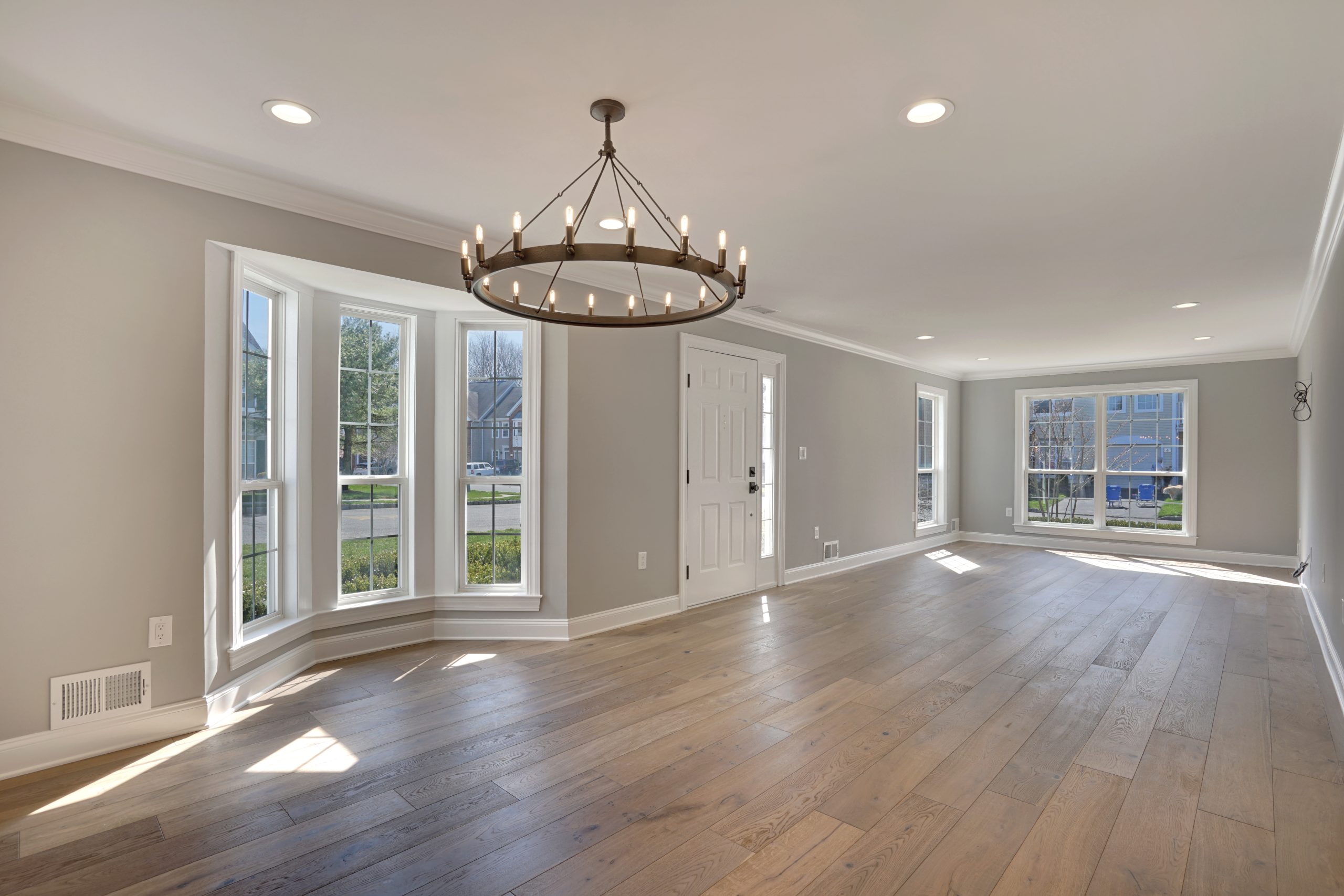 Bright, empty living room with large bay windows, light gray walls, recessed lighting, and wooden floors. A round chandelier hangs from the ceiling, and sunlight streams in through the windows.