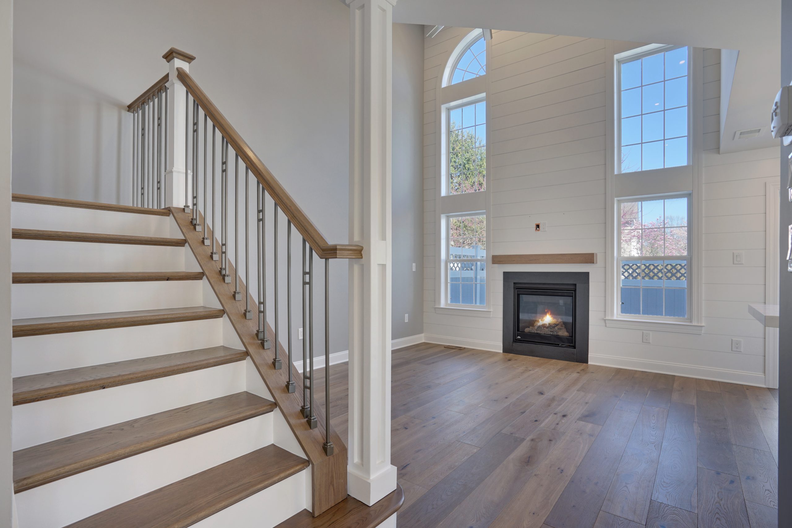 Modern living room with large windows, vaulted ceiling, and light wood floors; a staircase with wood steps and metal railings leads upstairs. A fireplace is built into a shiplap accent wall.