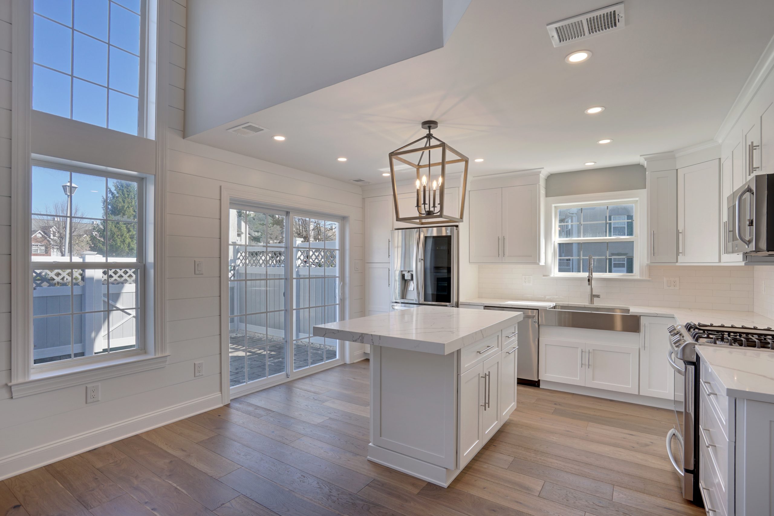 Modern, bright kitchen with white cabinets, marble island, stainless steel appliances, farmhouse sink, wood floors, large windows, and sliding glass doors leading to a fenced patio. Black lantern pendant light hangs over the island.