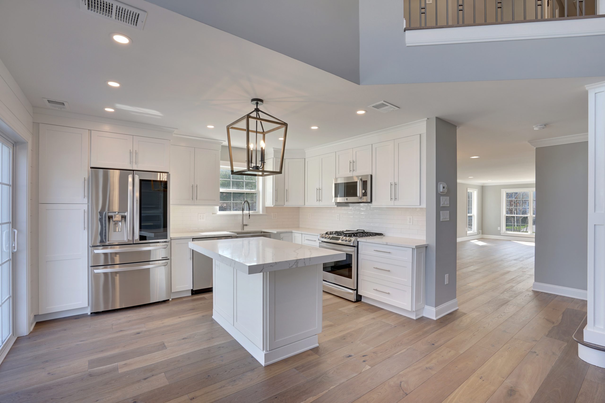 Modern kitchen with white cabinets, stainless steel appliances, a central island with marble countertop, light wood flooring, and open layout leading to a spacious living area with large windows.