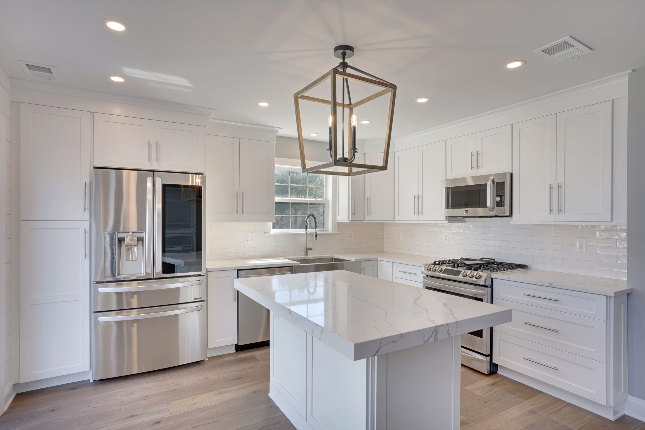 Modern kitchen with white cabinets, stainless steel appliances, a large central island with a marble countertop, a geometric pendant light, subway tile backsplash, and light wood flooring.