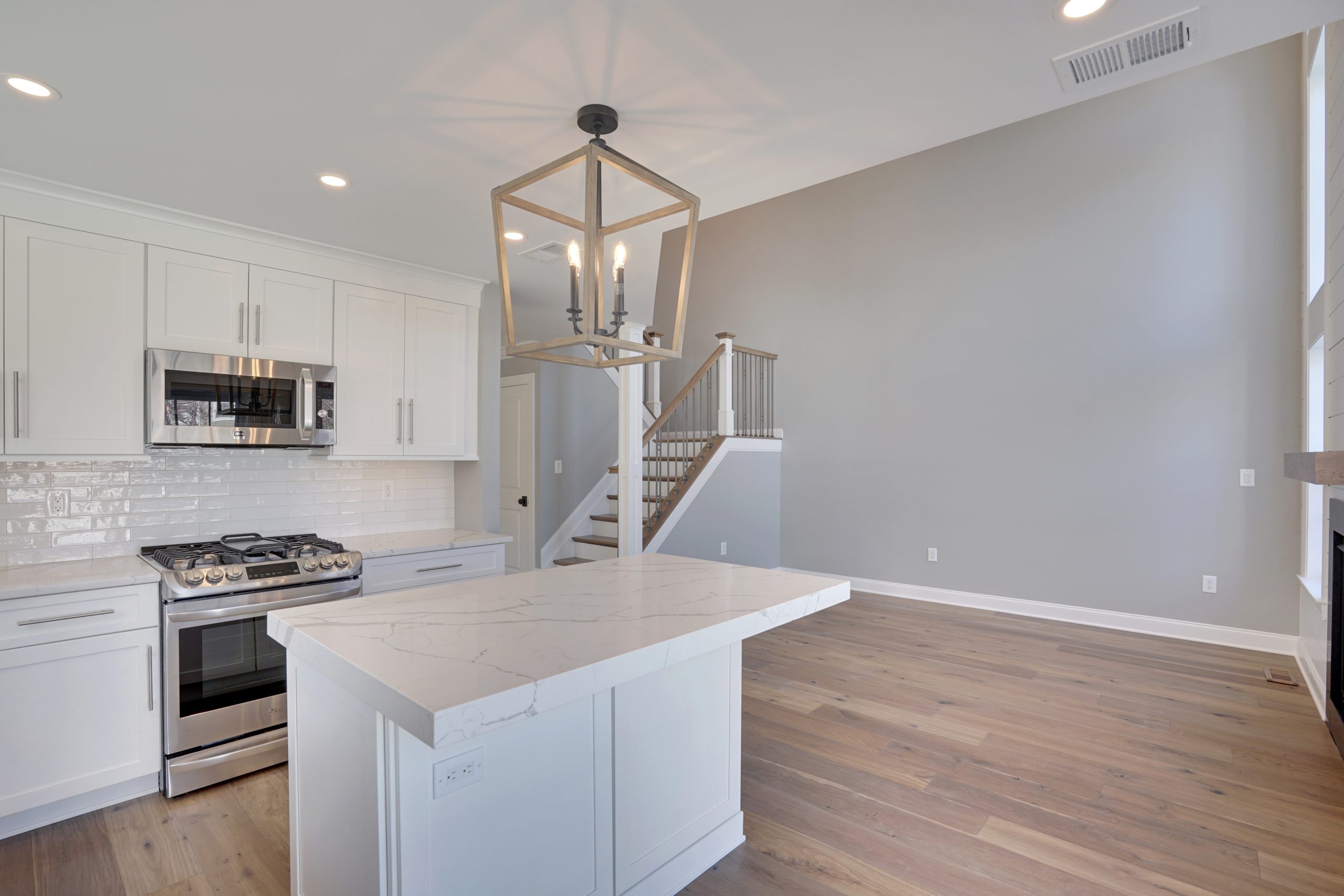 Modern kitchen with white cabinets, stainless steel appliances, a marble island, light hardwood floors, a geometric pendant light, and stairs leading to an upper level against a light gray wall.