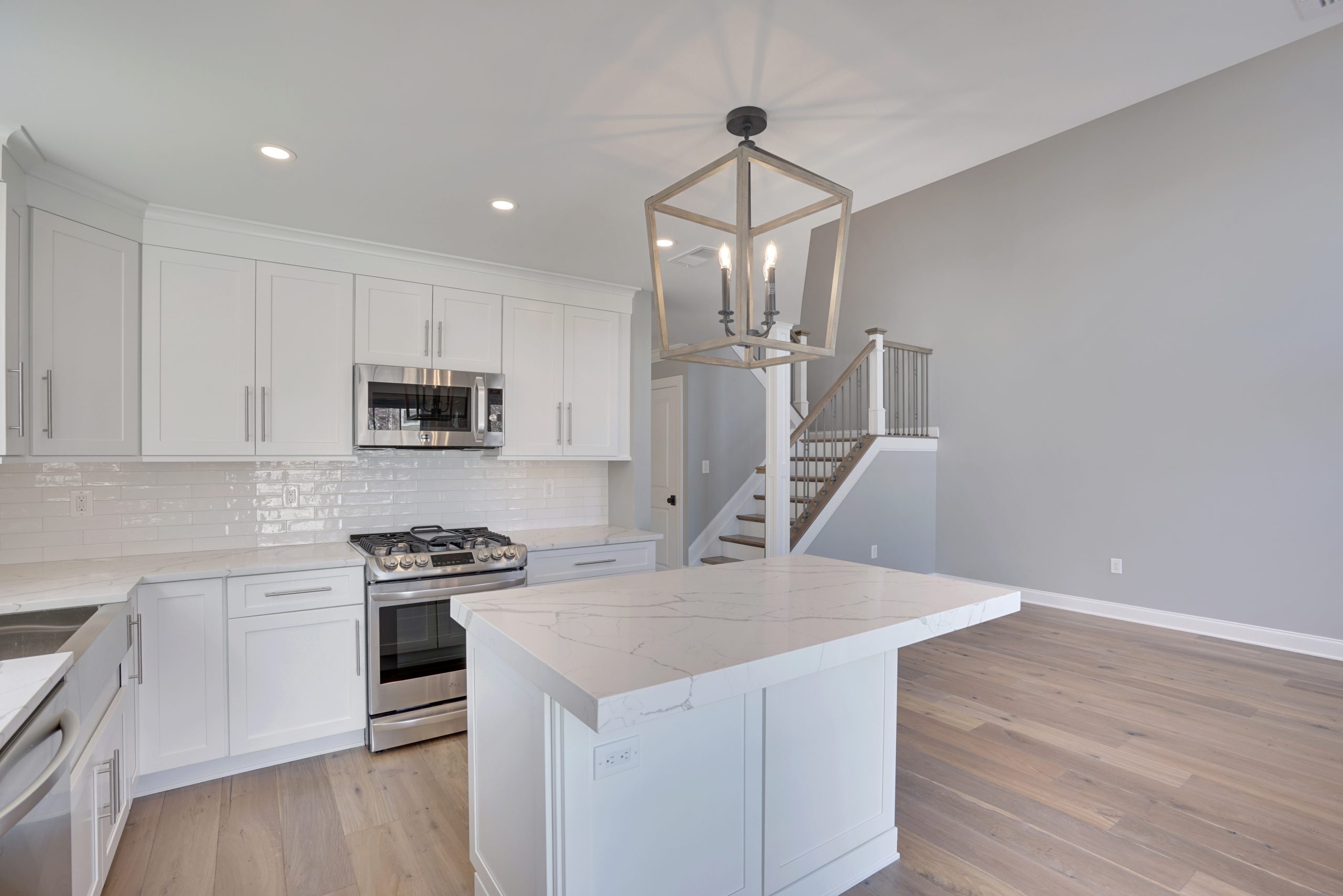 Modern kitchen with white cabinets, stainless steel appliances, marble island, light wood flooring, a geometric pendant light, and stairs with a metal railing against a light gray wall.
